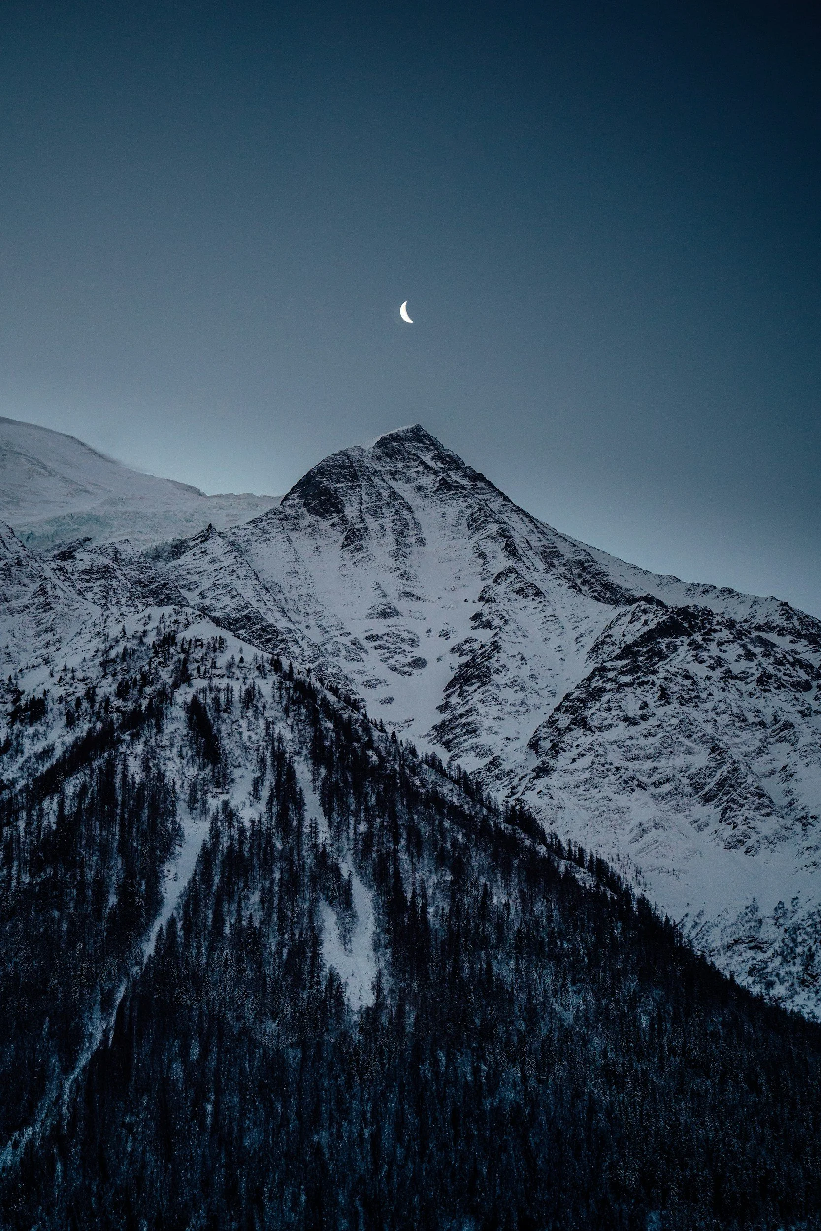 Snow-covered mountain peak under a clear blue sky with a crescent moon.