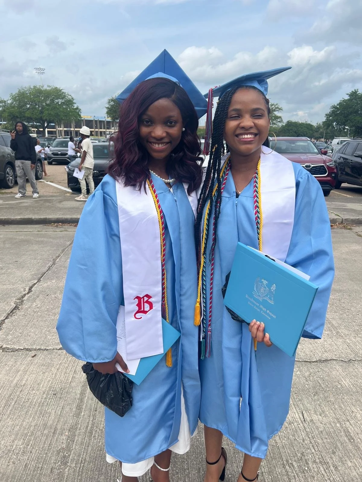 Two young women in light blue graduation gowns and caps, smiling at the camera, standing outside in a lot with other people and cars in the background.