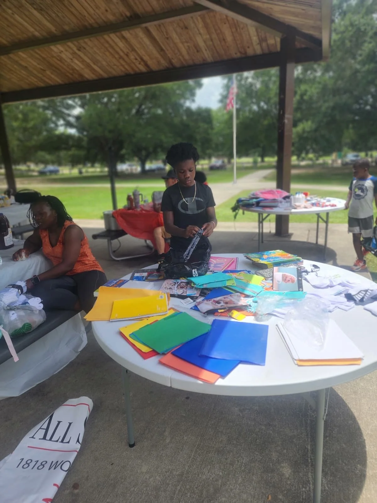 Children assembling and organizing school supplies at an outdoor event under a pavilion, with tables filled with notebooks, folders, and various school items.