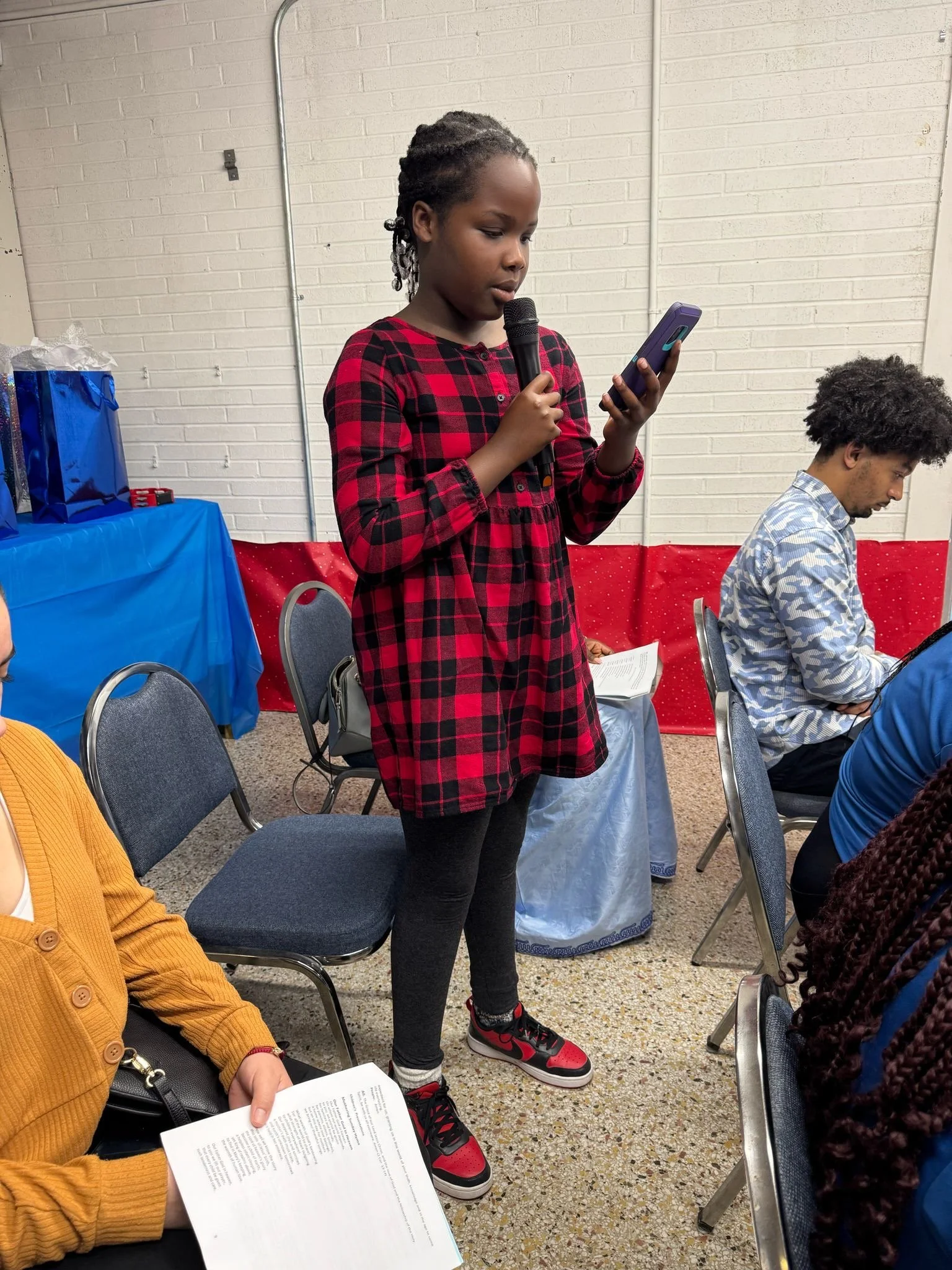 A young girl with braids in a red and black plaid dress holds a microphone and looks at her phone while standing among seated people at an indoor event.