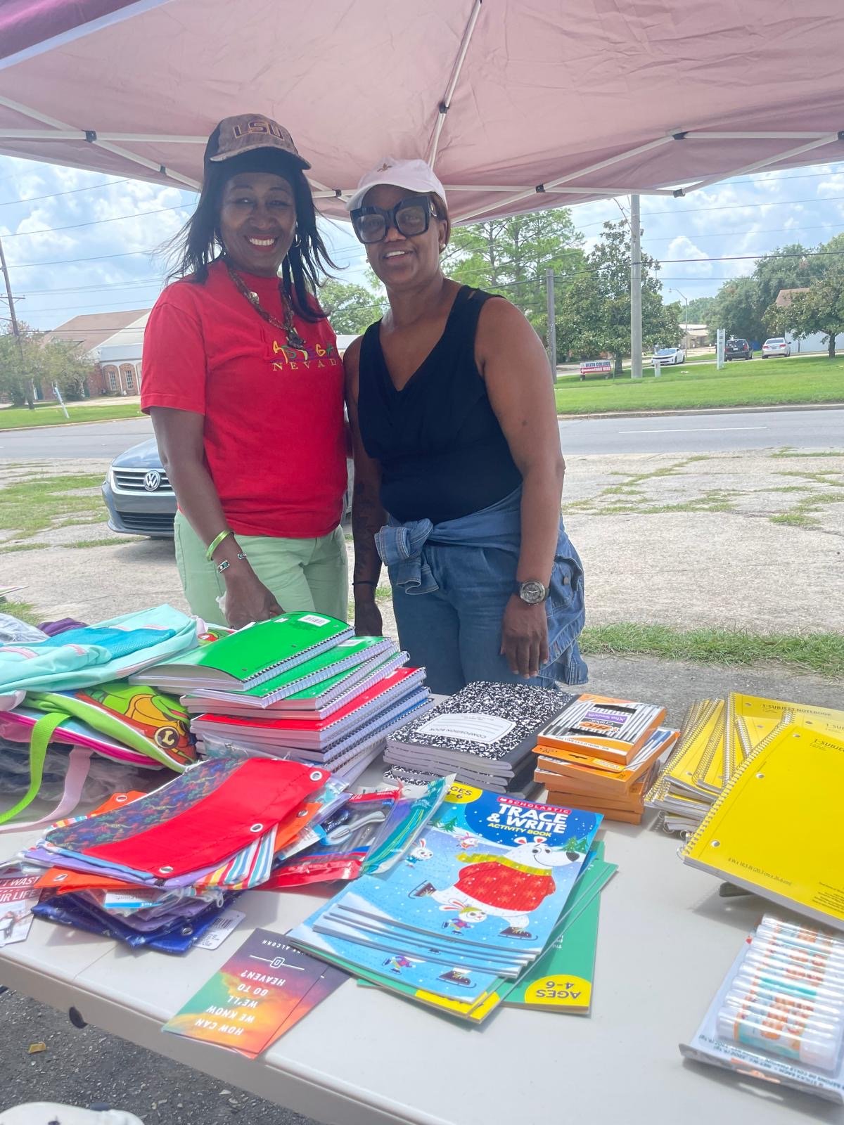 Two women standing behind a table filled with school supplies, under a pink canopy outdoors on a bright day.