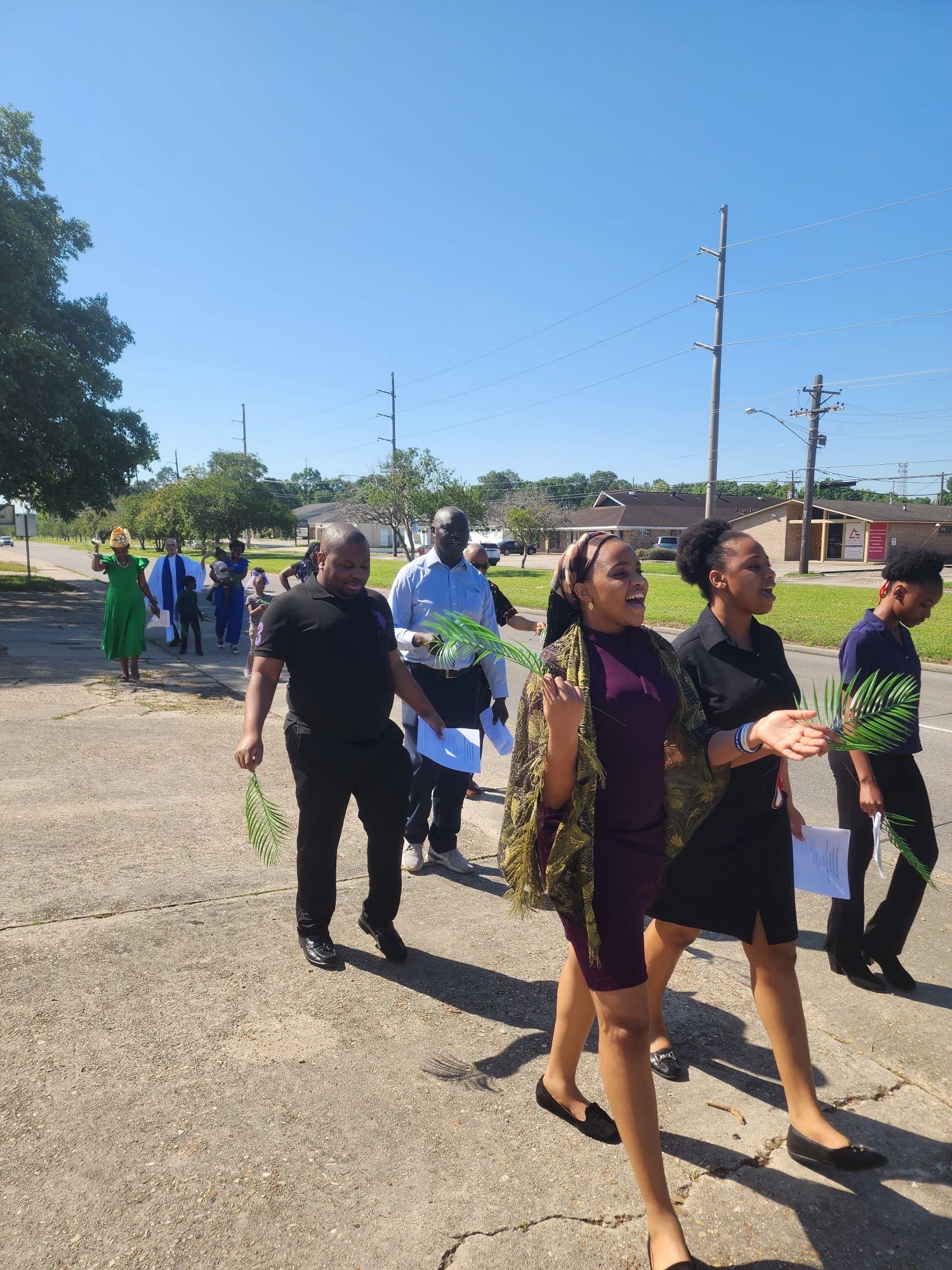 Group of people walking outdoors on a sunny day, some holding green palm leaves and papers, smiling and talking.