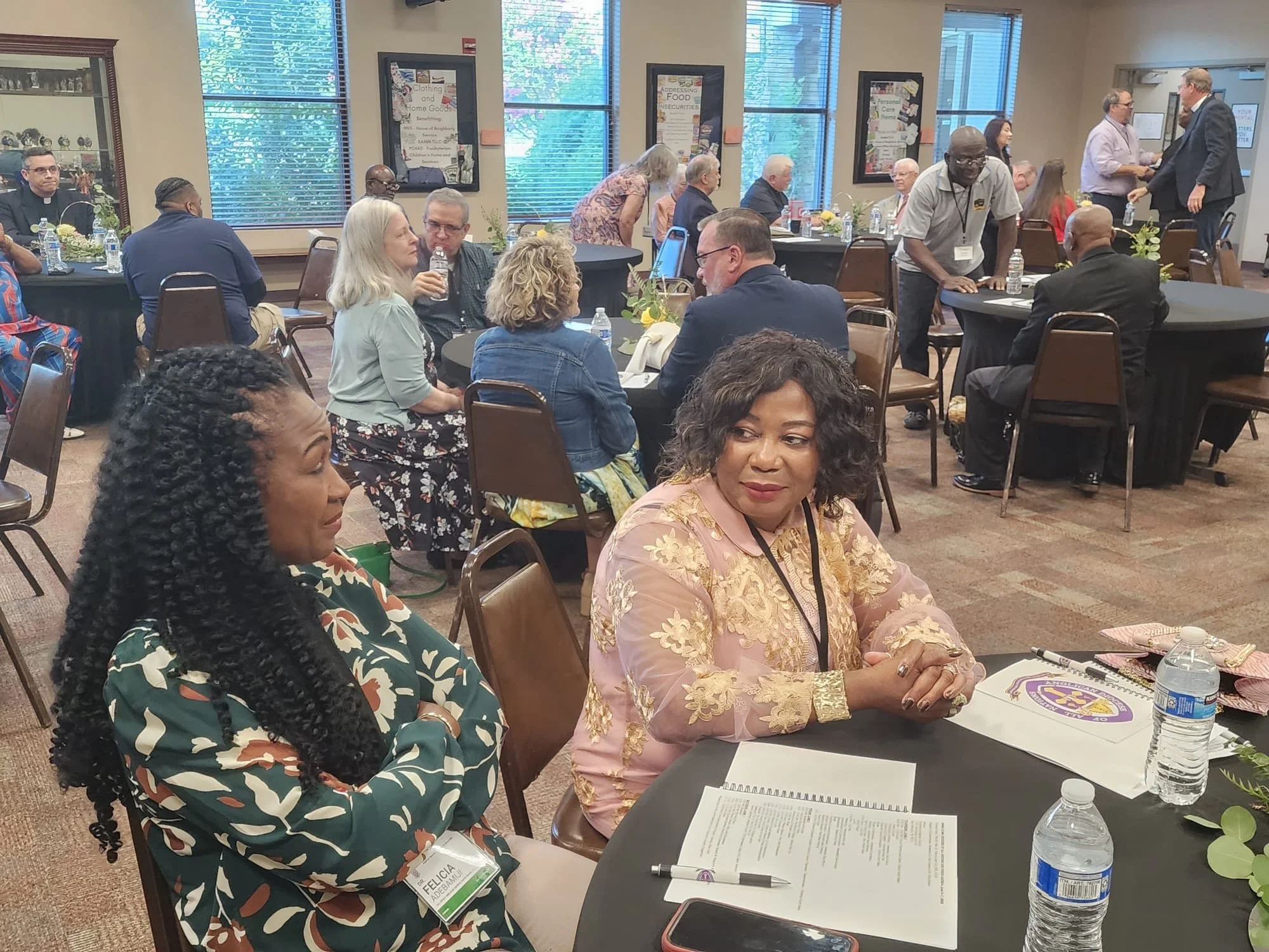 Conference room with diverse attendees sitting at round tables, some conversing, others listening, with papers, water bottles, and conference materials on the tables, and posters on the walls.