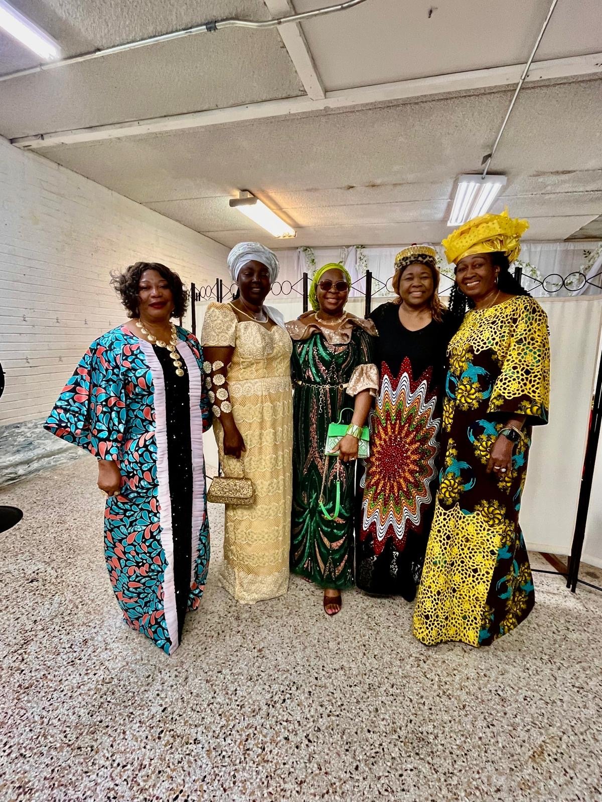 Five women dressed in colorful traditional African attire, standing together and smiling for a photo indoors.