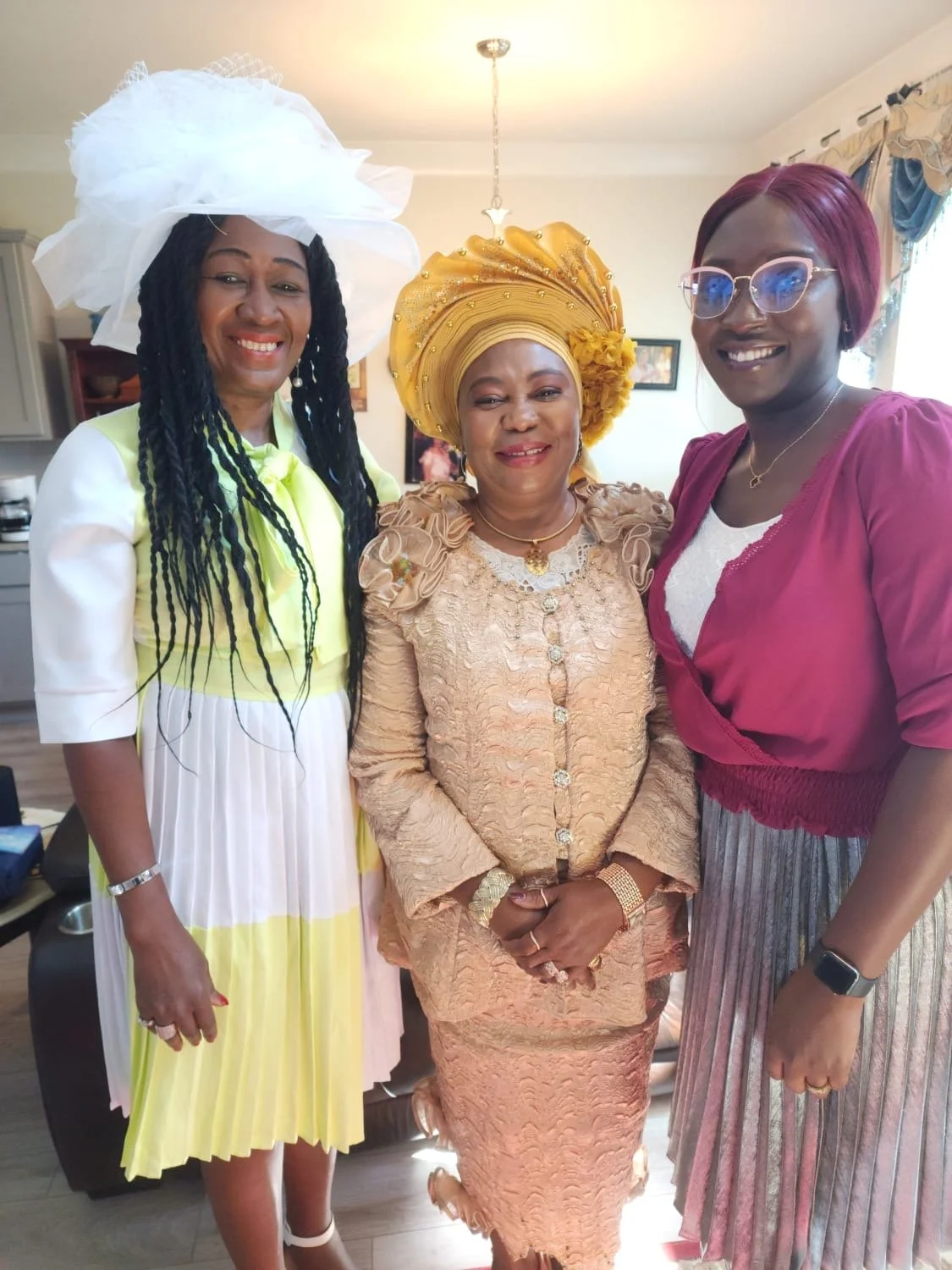 Three women smiling and dressed in colorful dresses and headpieces, standing indoors in a living room.