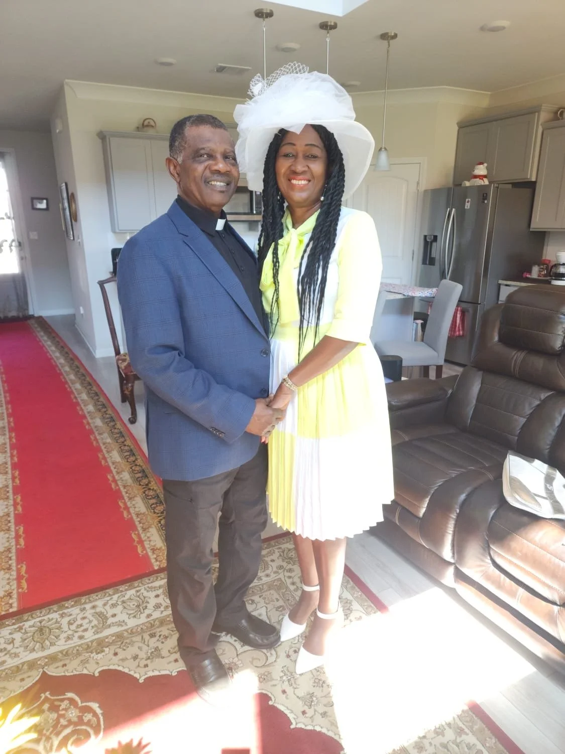 A man and a woman standing together inside a home, holding hands and smiling. The man is wearing a blue blazer and a clerical collar, and the woman is dressed in a yellow and white dress with a large white hat.