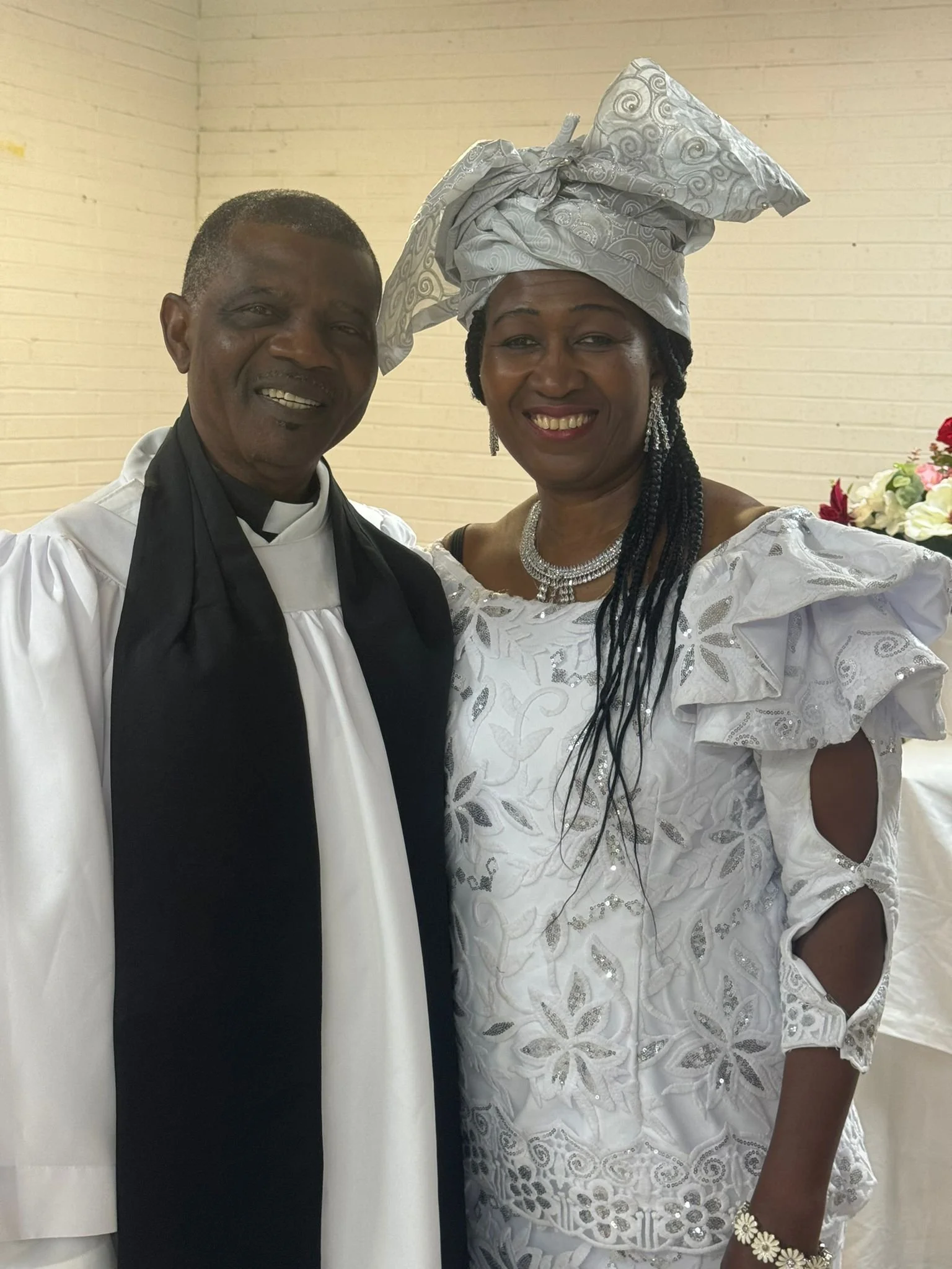 A smiling man and woman dressed in white with intricate lace and embroidery, standing close together indoors against a beige brick wall. The woman is wearing a large matching headwrap and jewelry.