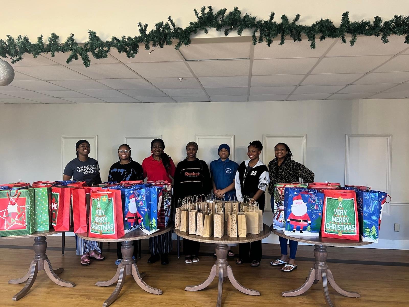Seven women are standing behind three tables filled with Christmas-themed gift bags and paper bags. The women are smiling, and the room is decorated with a green garland hanging from the ceiling.
