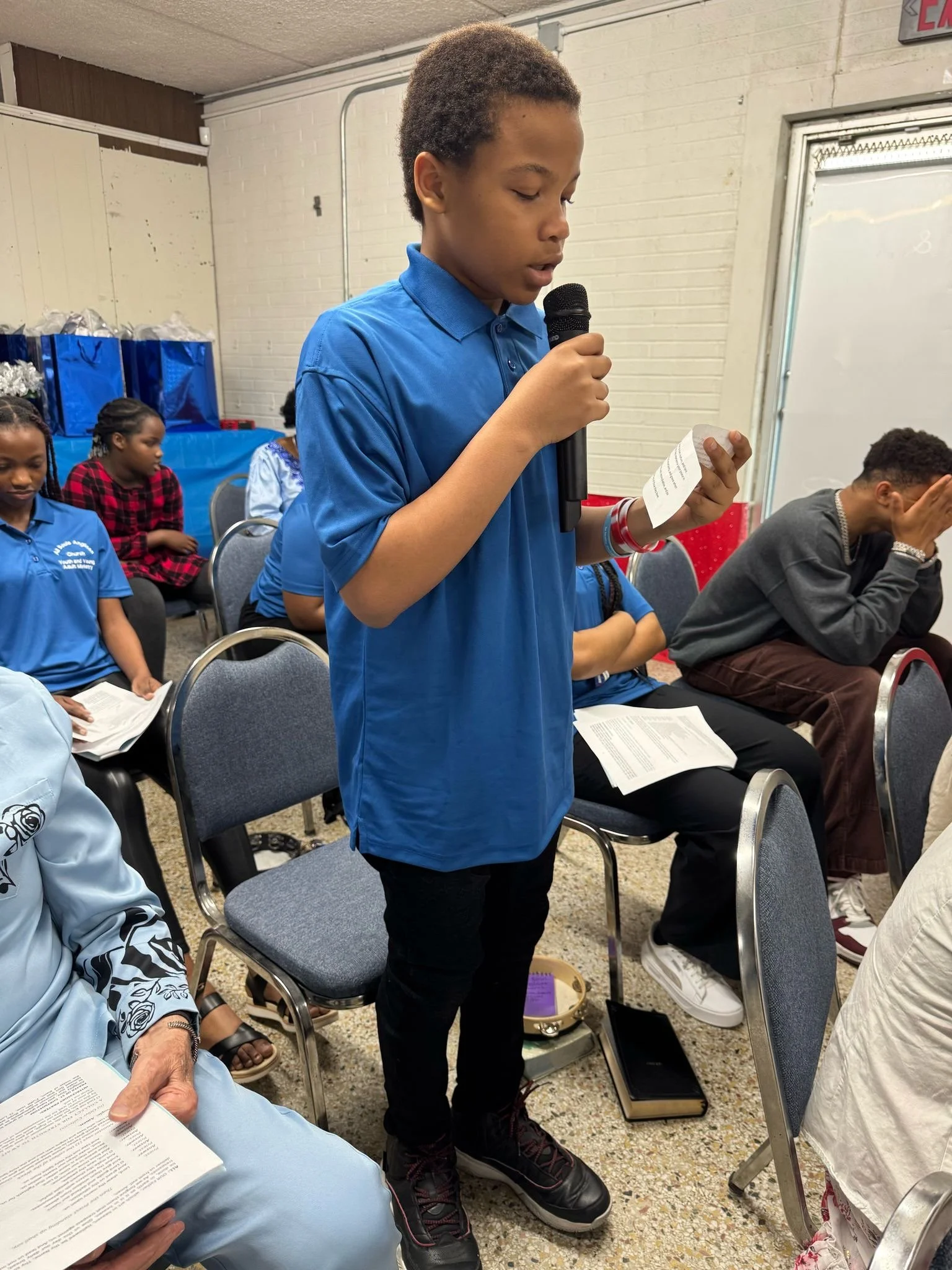 A young boy with curly hair wearing a blue polo shirt and black pants is standing and reading from a paper while holding a microphone during a group meeting or event. Several people are sitting around him, some reading papers and others appearing to be praying or listening, in a room with white brick walls and a window.