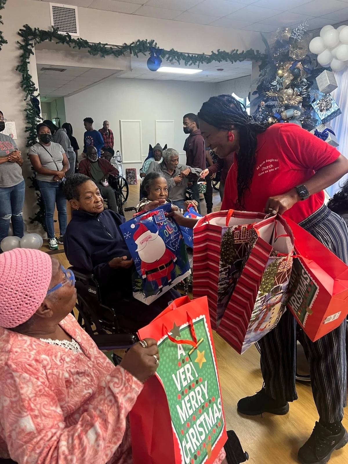 A woman in a red shirt handing out Christmas gifts to elderly women seated in a room decorated with a Christmas tree, balloons, and holiday decorations, during a holiday celebration.