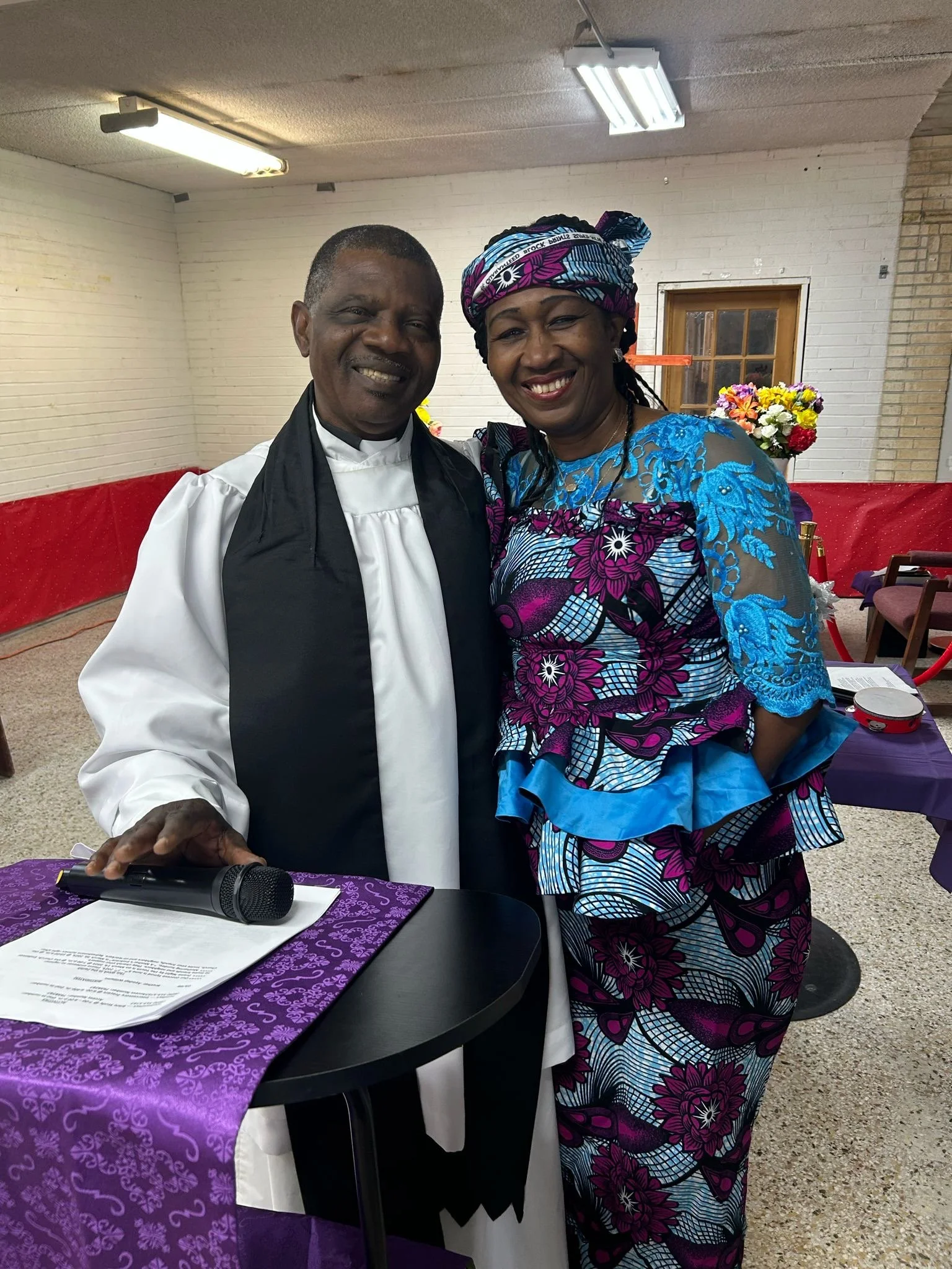 A smiling man in religious attire standing next to a smiling woman in colorful traditional clothing in an indoor setting, with flowers and tables in the background.