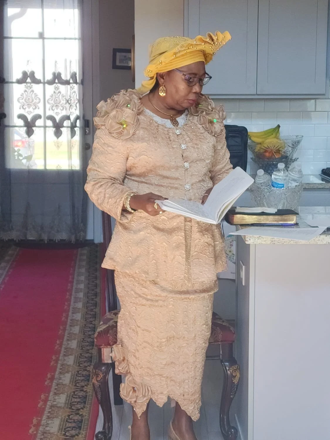 A woman dressed in beige lace traditional attire with ruffled details stands and reads a book in a kitchen with gray cabinets, a white countertop, and a fruit bowl.