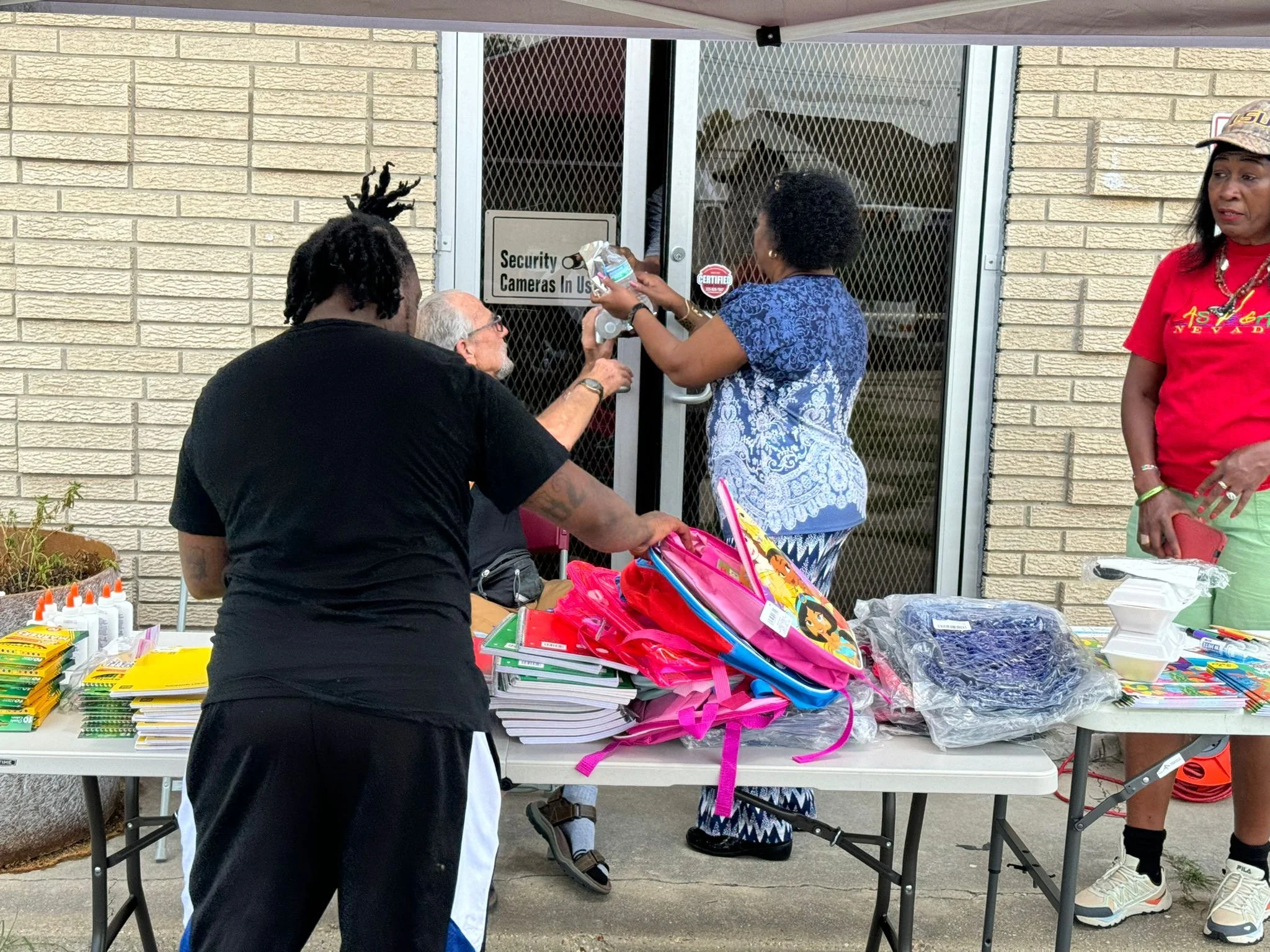 People standing and sitting at tables outside a building, with backpacks and school supplies on the tables, as a woman hands a package to someone through a glass door.