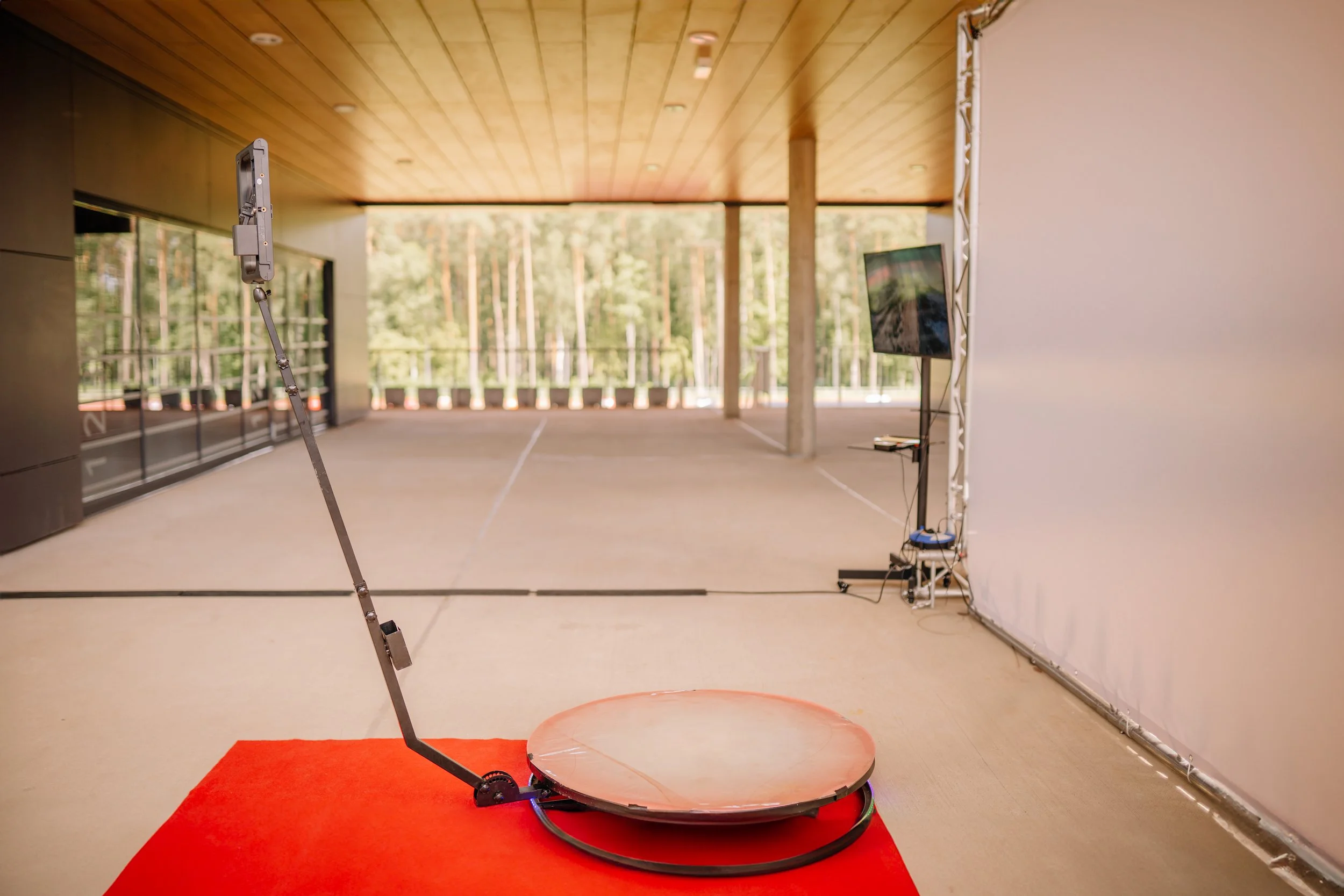 A virtual reality motion simulation device on a red mat in a modern, open indoor space with large windows, a wooden ceiling, and a view of trees outside. The device has a circular platform and a large mounted monitor.