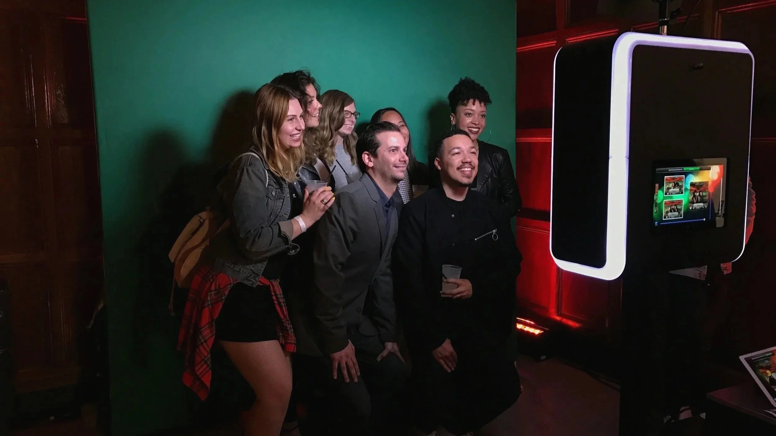 A group of six people, five women and two men, are gathered for a photo in front of a green screen at a photo booth with a digital screen and a large mirror on the side.