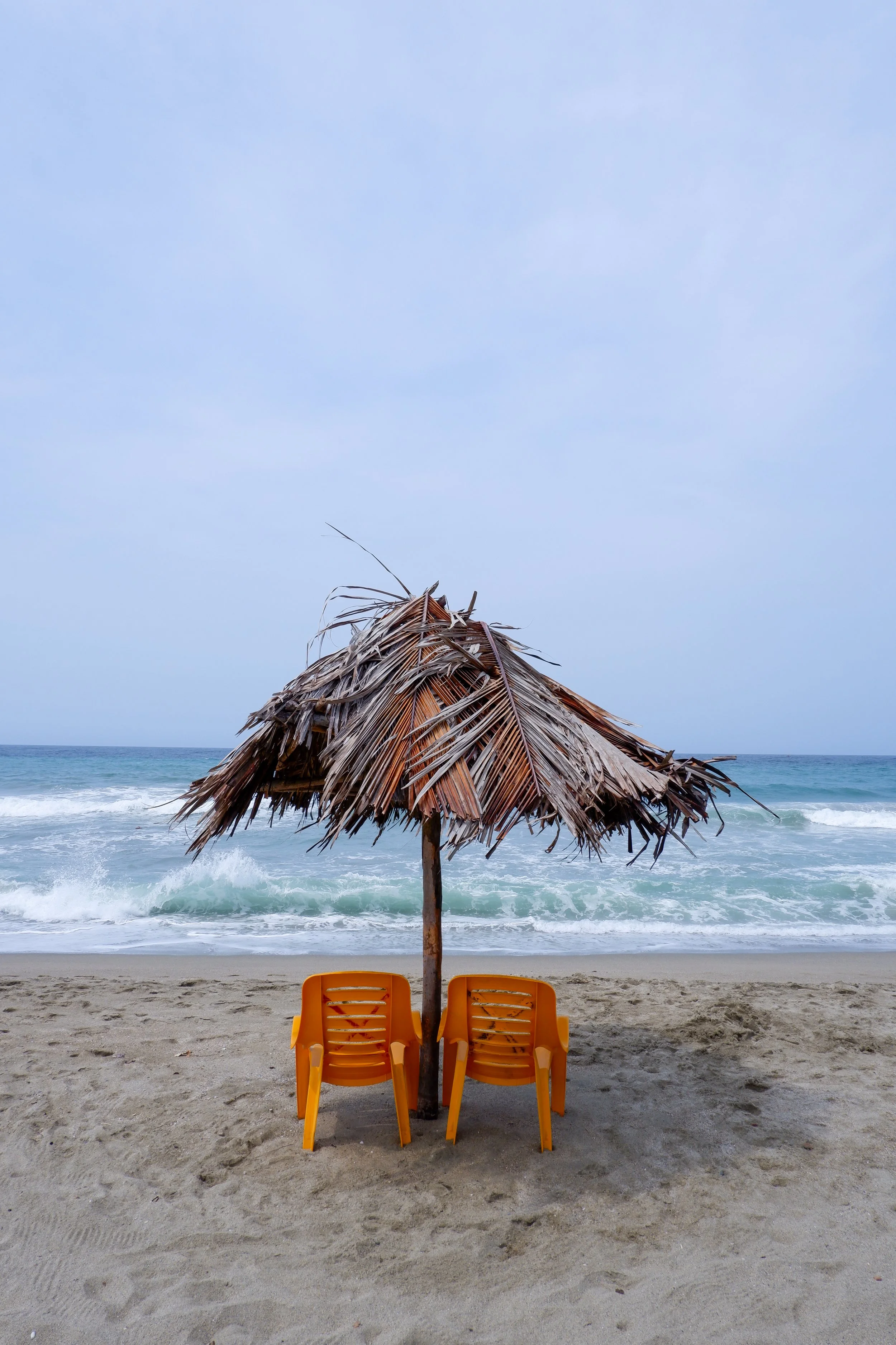 Empty beach with two orange chairs under a rustic palm leaf thatched umbrella facing the ocean with waves and a cloudy sky