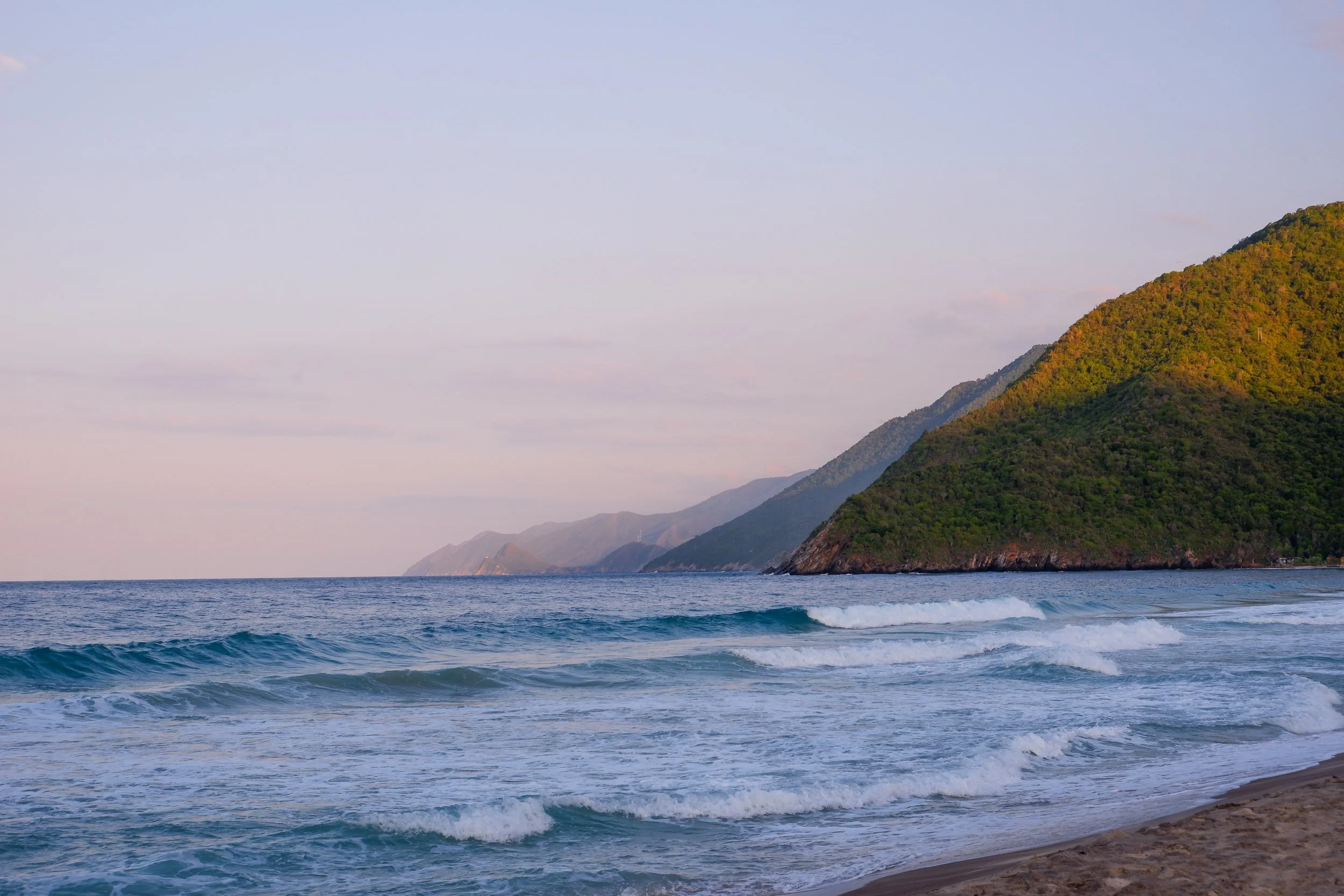 Beach with waves and green hills in the distance, under a pastel sky.