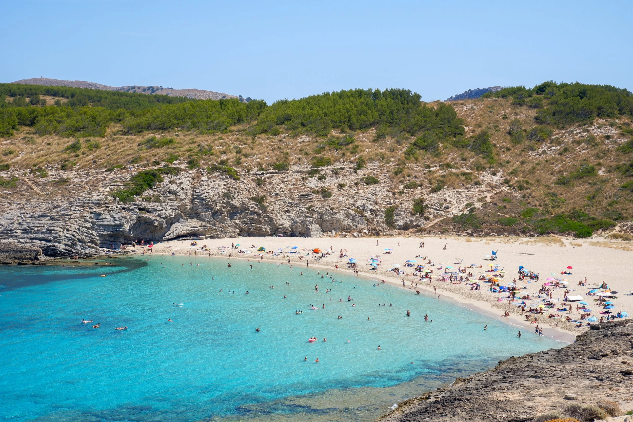 A sandy beach with numerous people, umbrellas, and swimming in turquoise water, surrounded by rocky cliffs and green hillside.