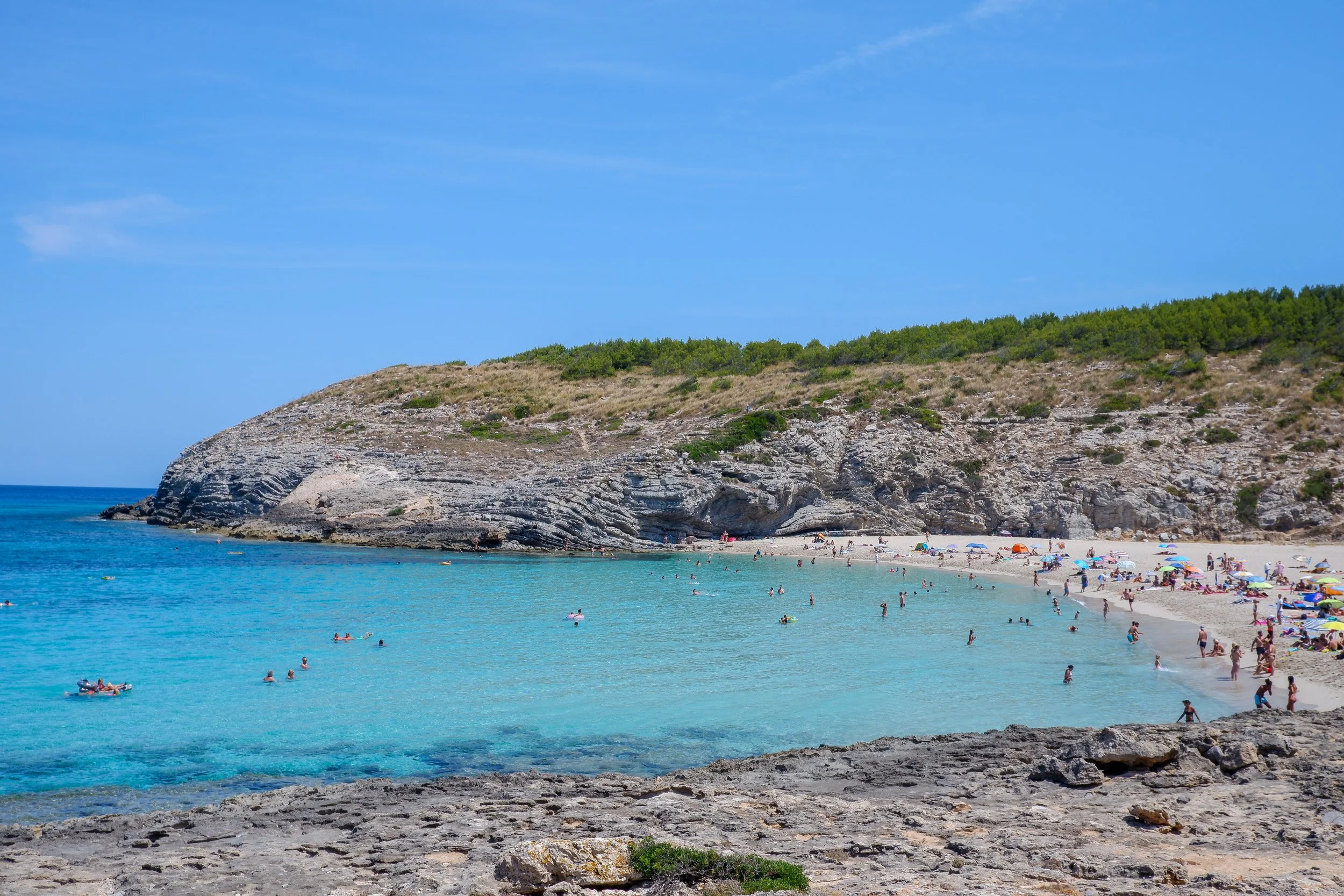 A scenic beach with turquoise water, sandy shore, rocky cliffs, and many people swimming, sunbathing, and relaxing under umbrellas on a bright sunny day.