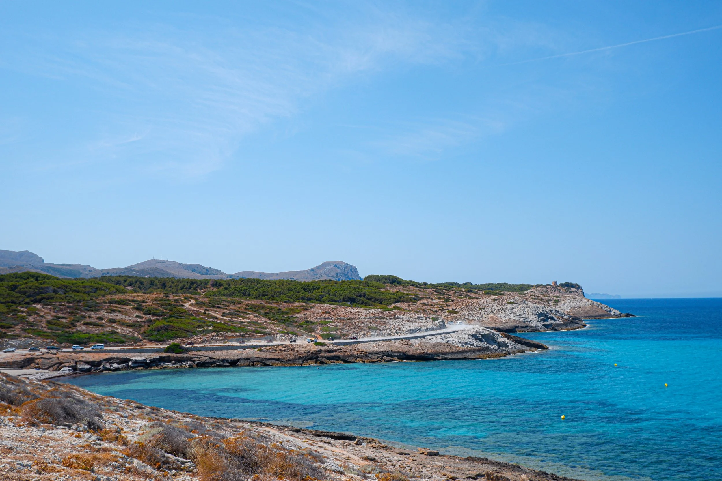 Coastal landscape with turquoise waters, rocky shoreline, and green hills under a blue sky.