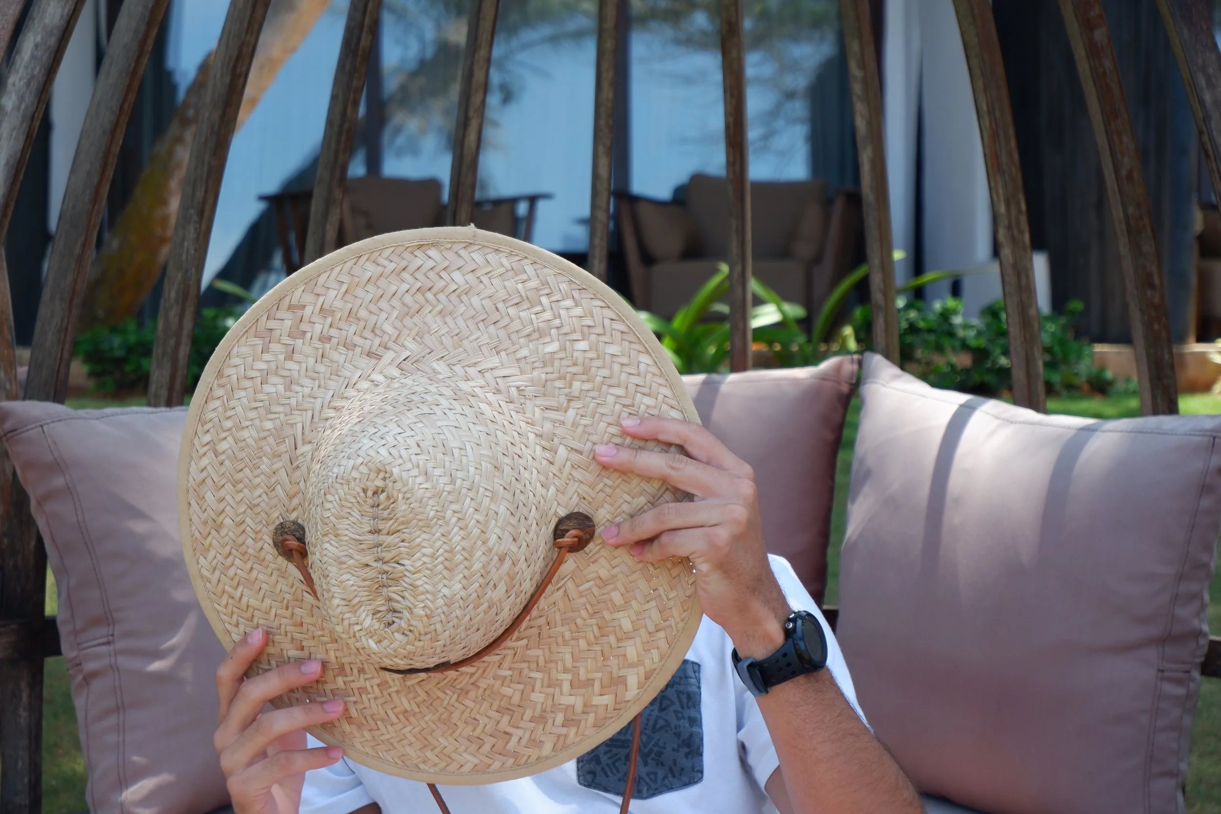 Person holding a straw hat covering their face while sitting on outdoor cushions in a garden or patio area.
