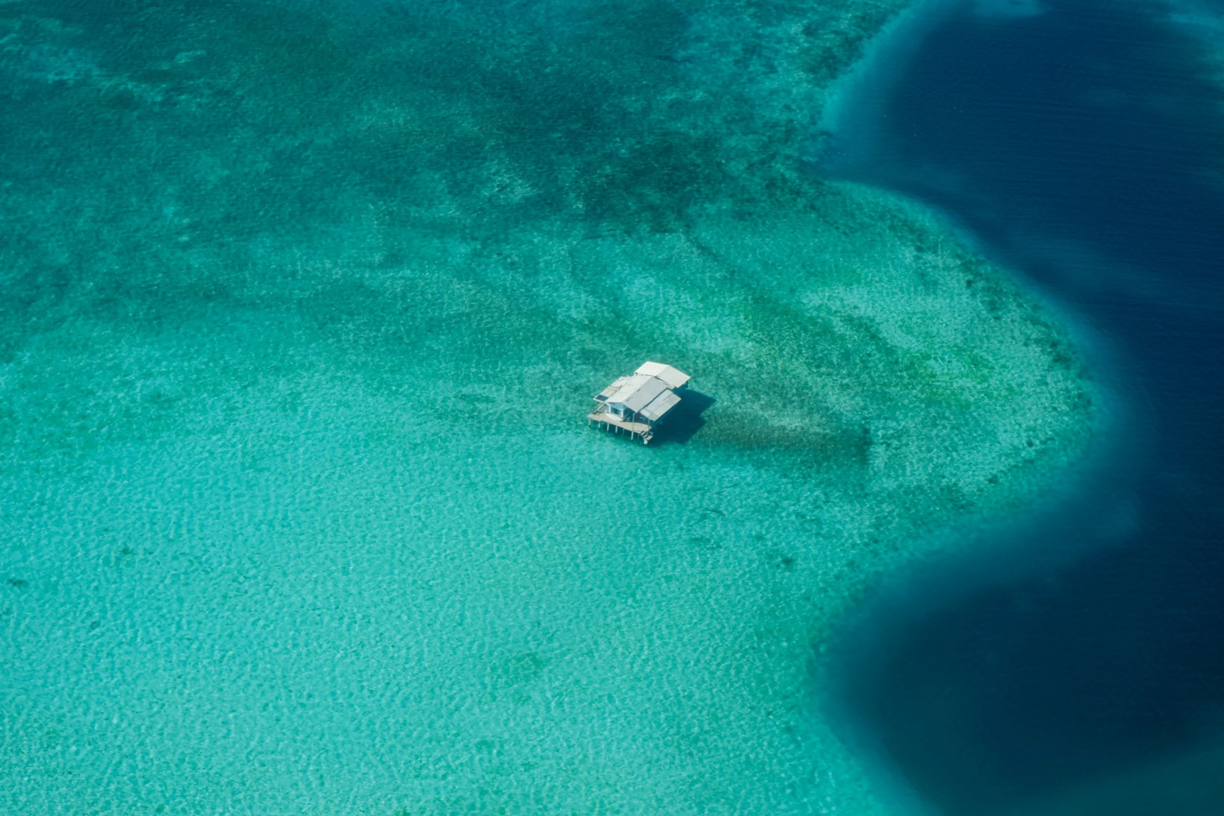 A small house on stilts surrounded by shallow turquoise water with clear coral reefs visible beneath the surface.