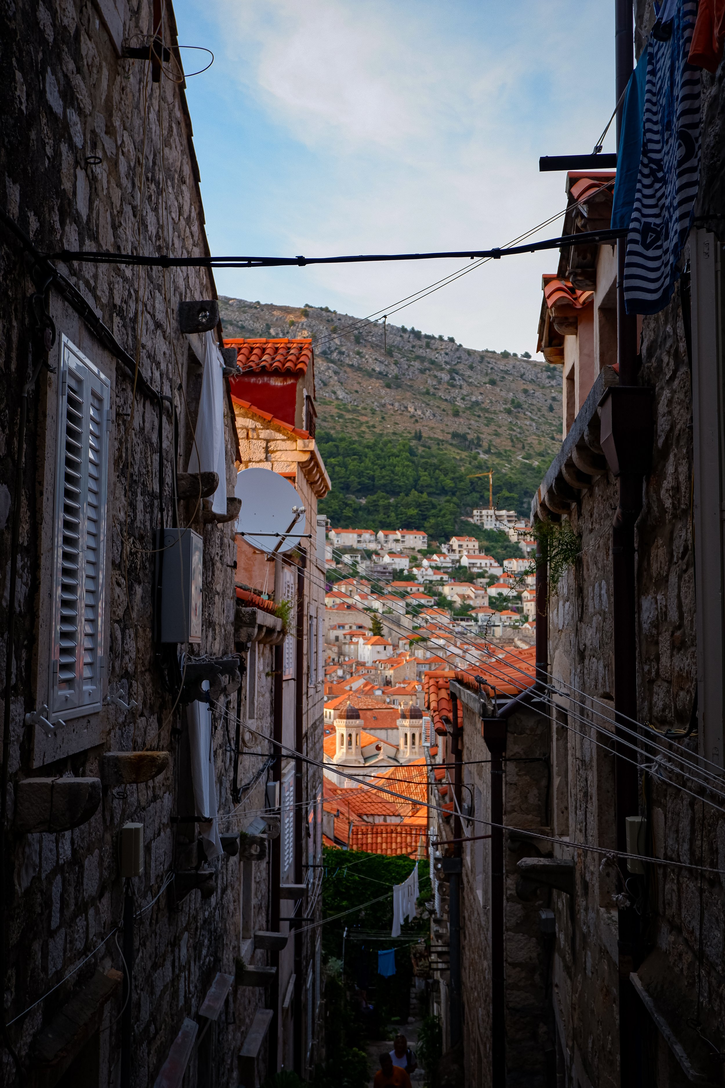 A narrow alleyway between stone buildings with laundry hanging on clotheslines, overlooking a hillside town with orange rooftops and green trees in the background.