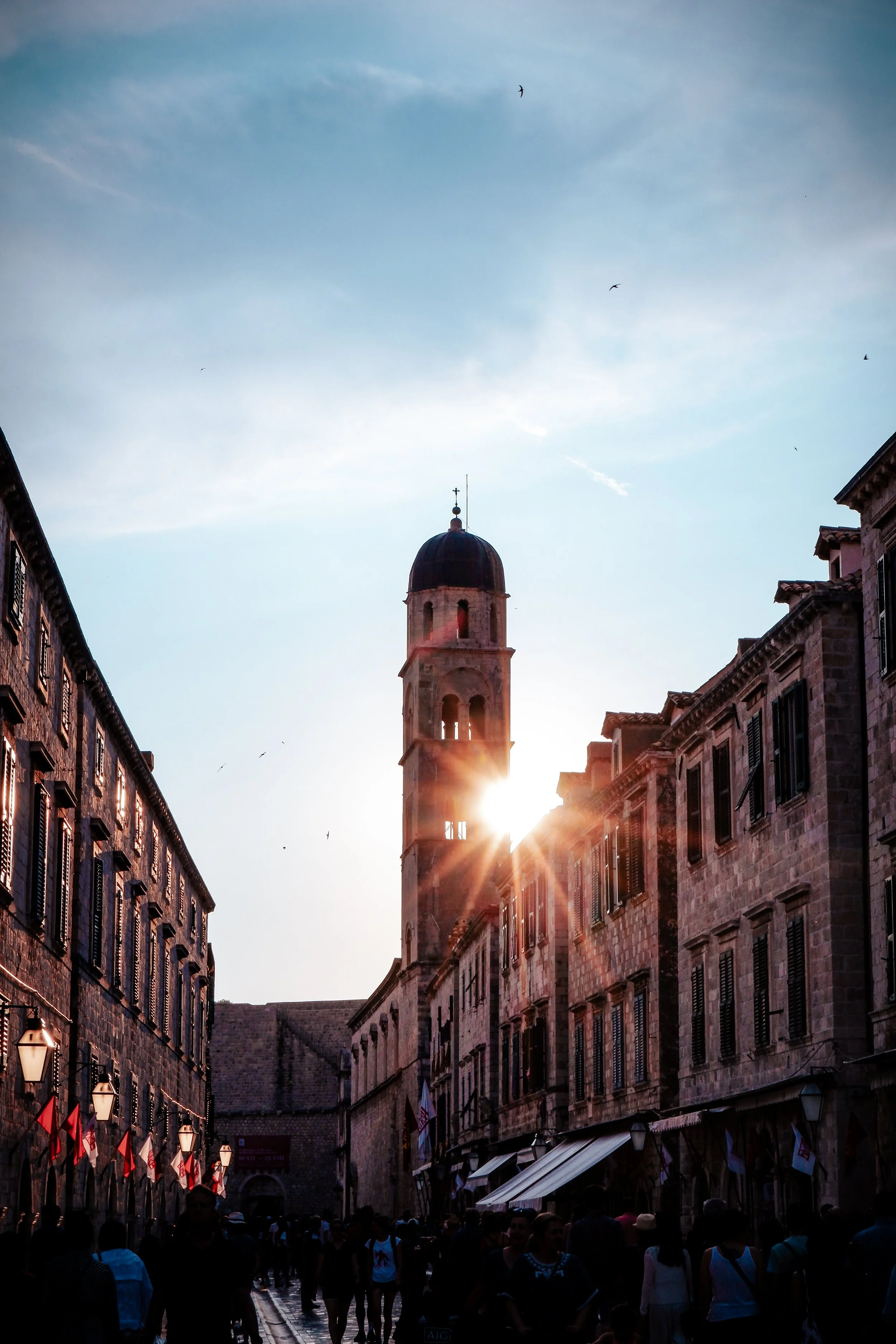 Sunset view of a historic European street with a clock tower in the background, crowds of people walking, buildings with shutters, and Flag banners along the street.