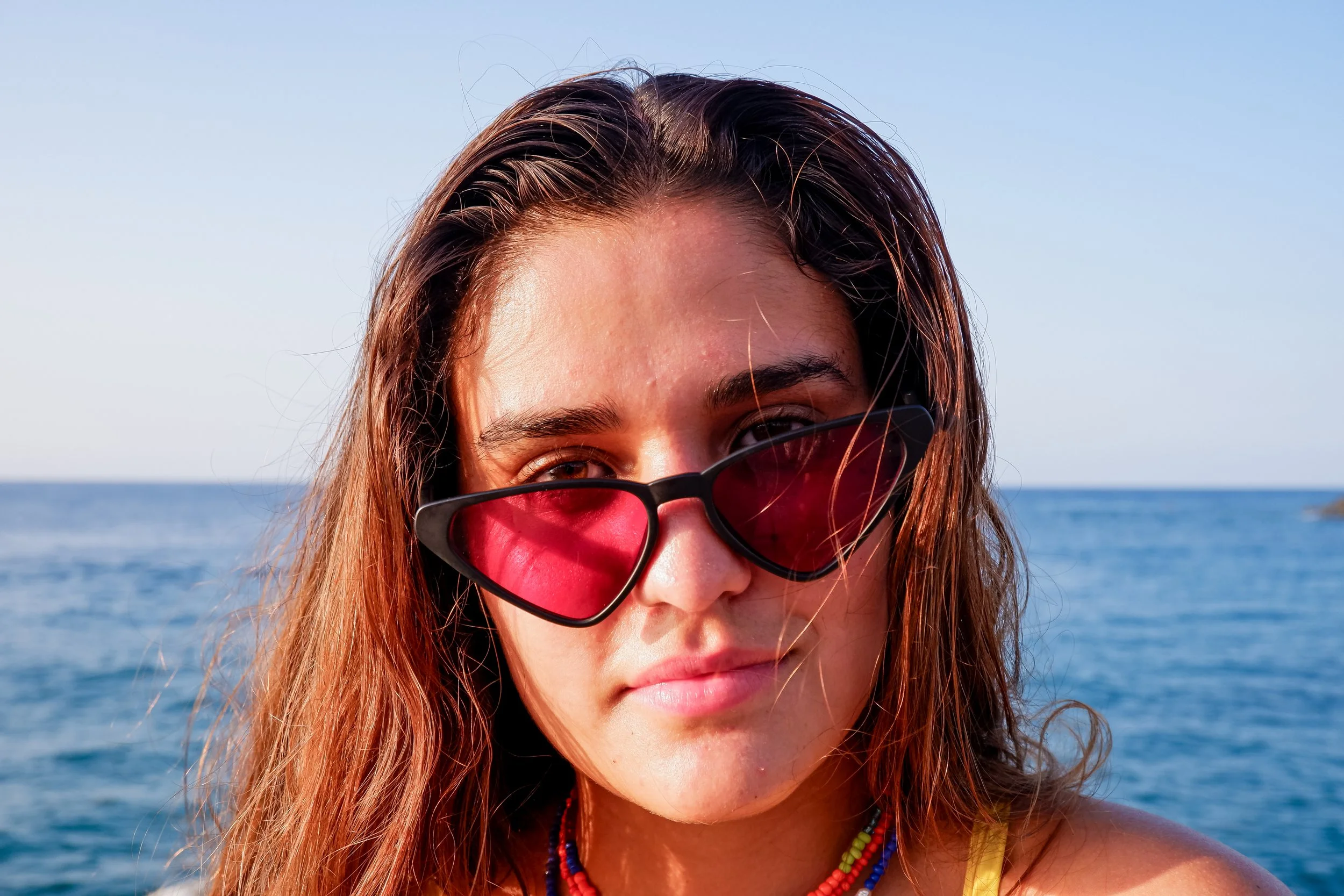 Woman with wet hair wearing red-tinted sunglasses near the ocean.