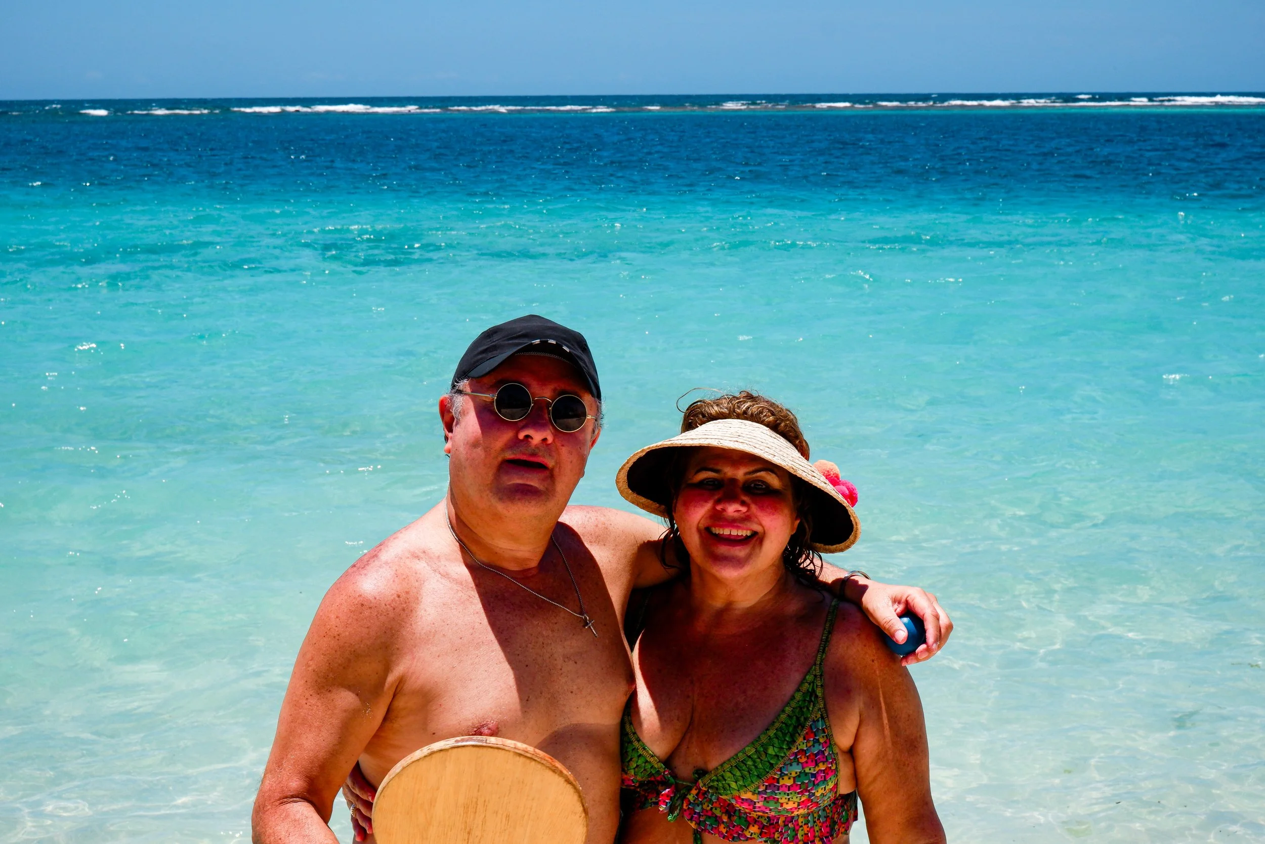 A man and woman standing in clear ocean water at the beach smiling, with the man wearing sunglasses and a black cap, and the woman wearing a sun hat and a colorful swimsuit.
