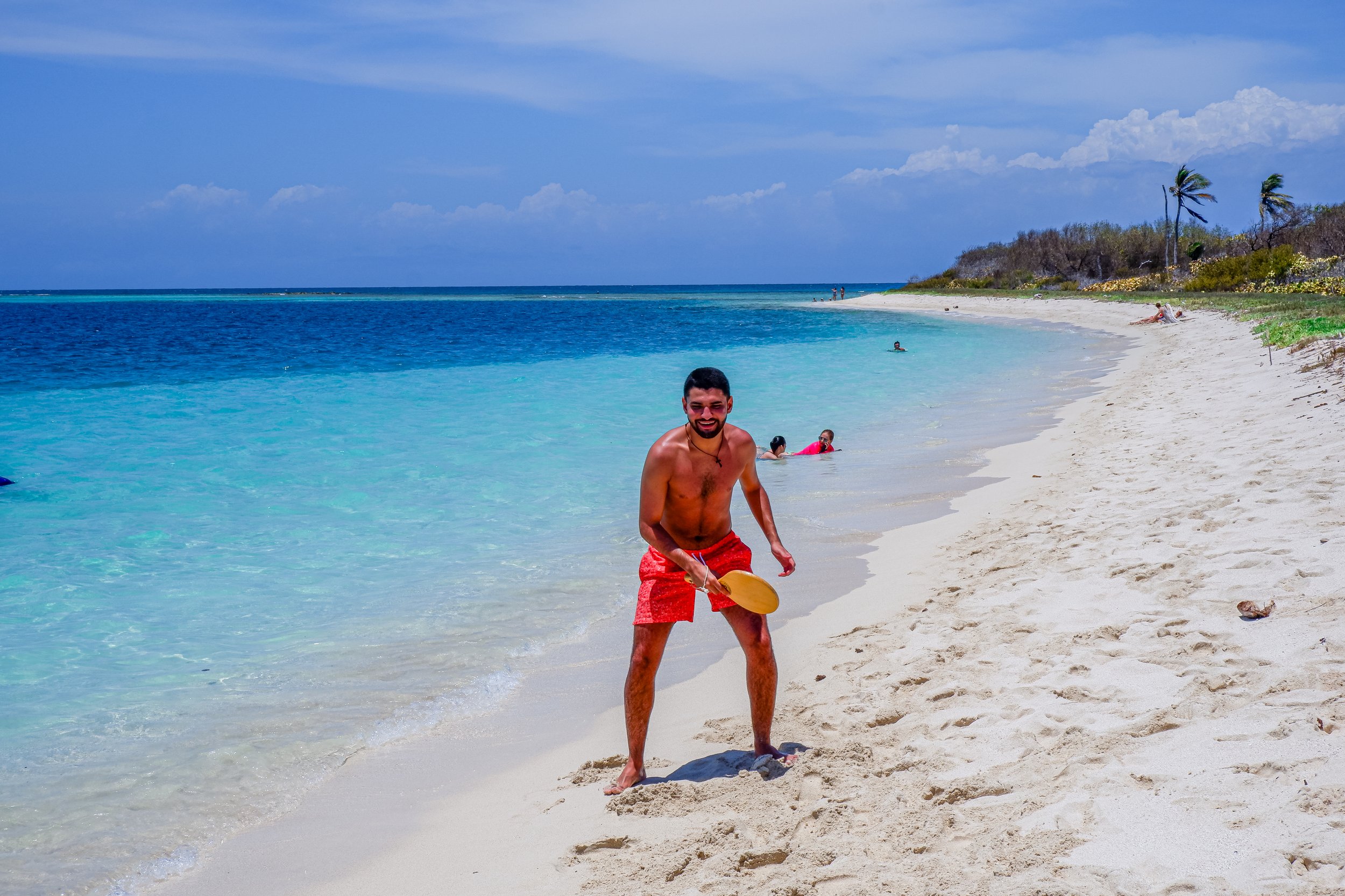 Smiling man in red swim shorts playing beach paddleball on sandy beach with turquoise water and a few people swimming, palm trees, and a partly cloudy sky in the background.