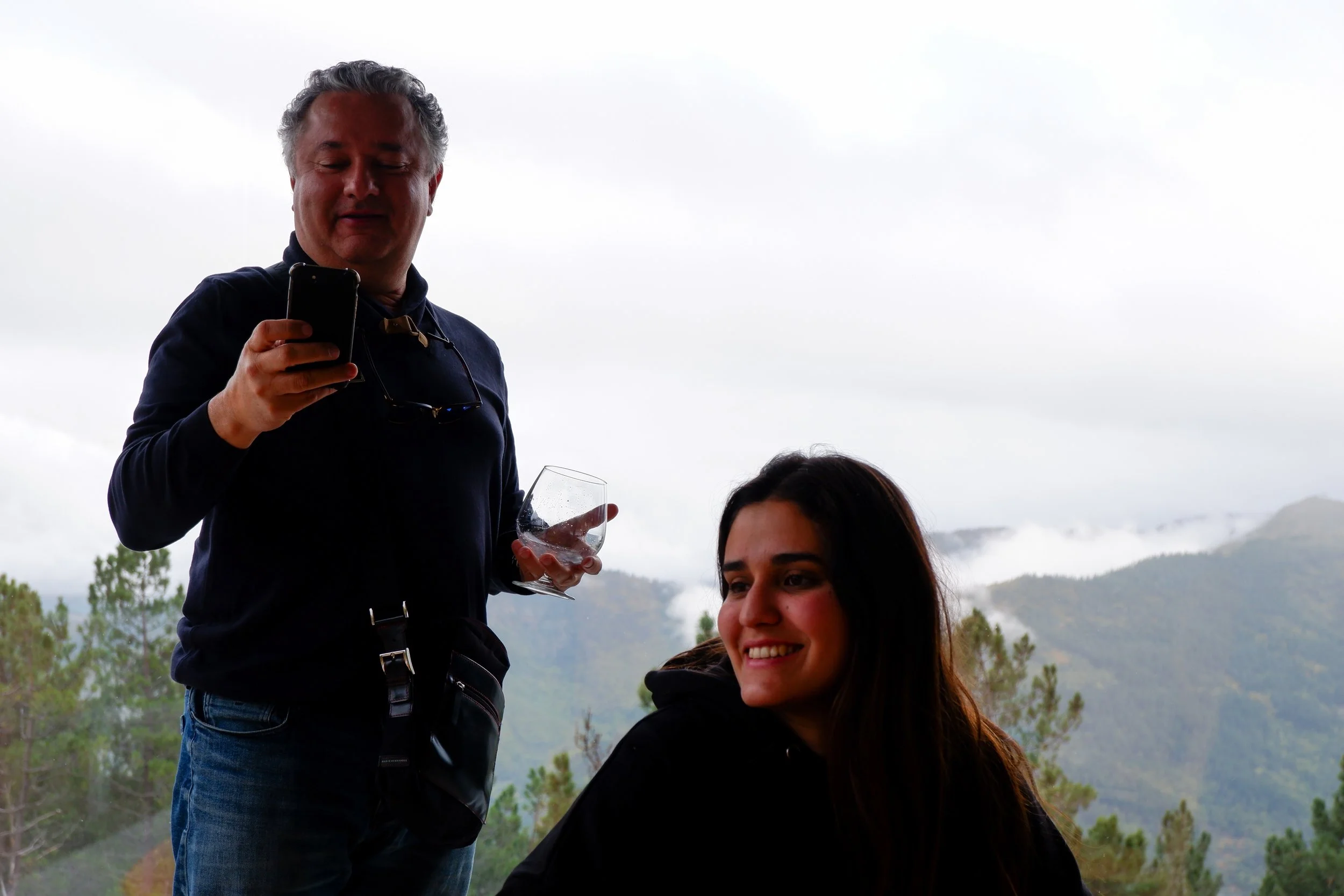 A man is looking at his phone with sunglasses hanging around his neck, holding a glass, while a woman sits nearby smiling. They are outdoors with a mountain and cloudy sky in the background.