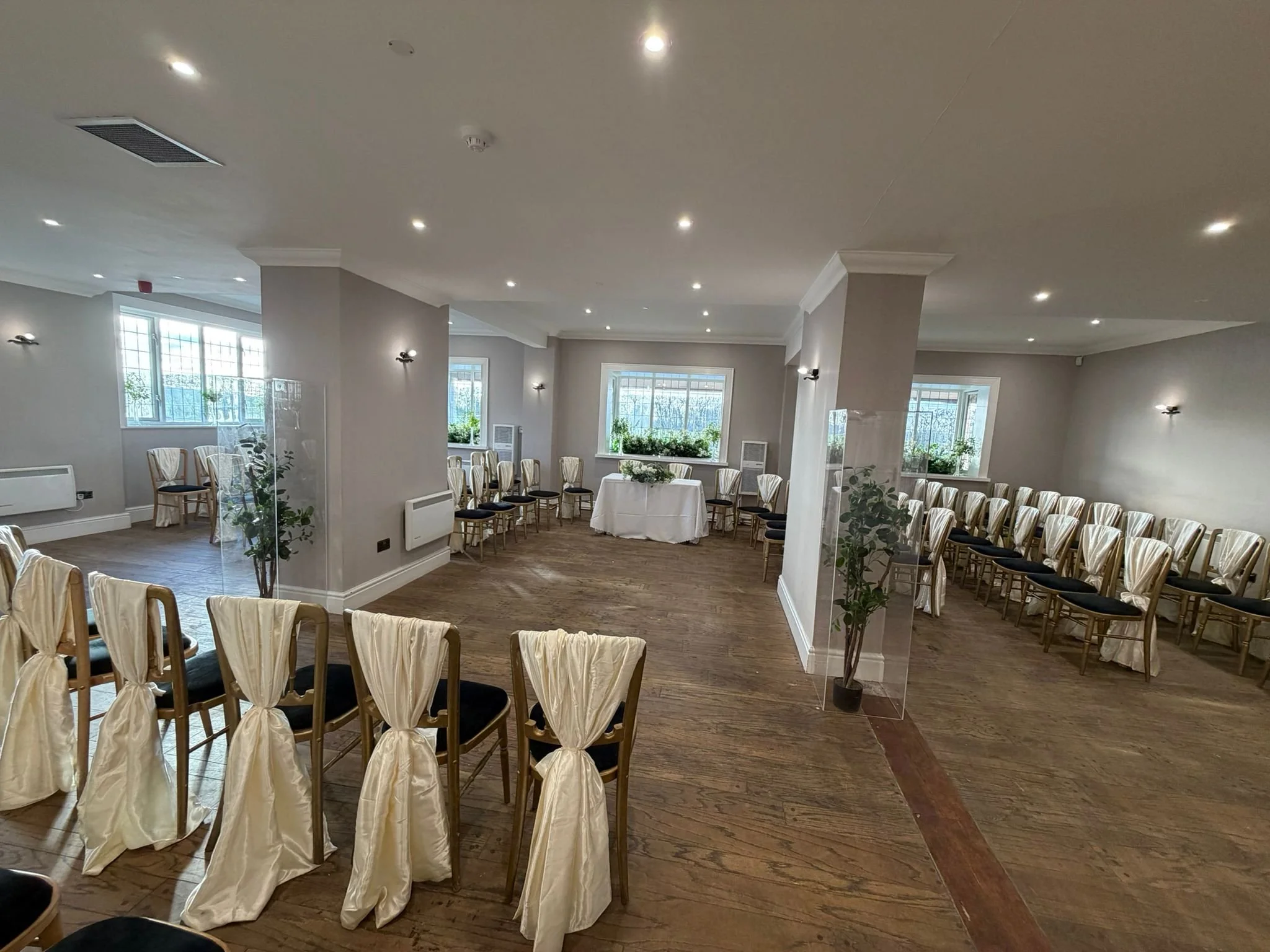 A spacious banquet room set up for a wedding or event with rows of chairs decorated with cream-colored fabric and black cushions, arranged along the walls and around a small table with a white tablecloth, large windows with natural light, and potted 
