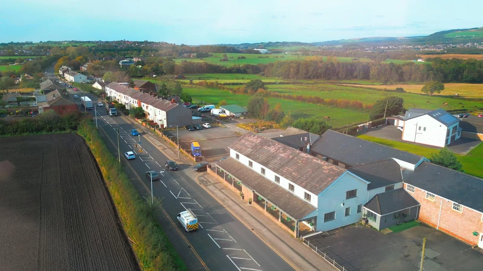 Aerial view of a small town with houses, parking lots, and a main road, surrounded by green fields and distant hills under a blue sky.