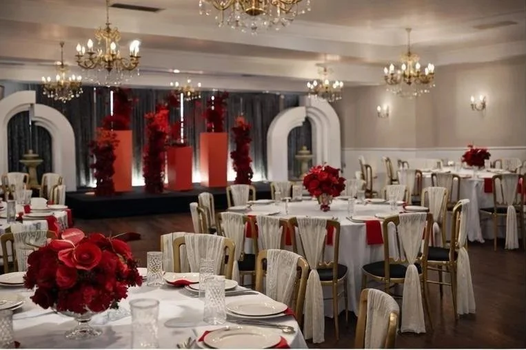 Elegant banquet hall decorated for a celebration with round tables covered in white tablecloths, red floral centerpieces, and chairs with white slipcovers and red accents, featuring a stage with red flower arrangements and a chandelier-lit ceiling.