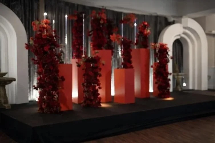 Stage decorated with red floral arrangements on tall stands and a backdrop with metallic streamers, set for an event or celebration.