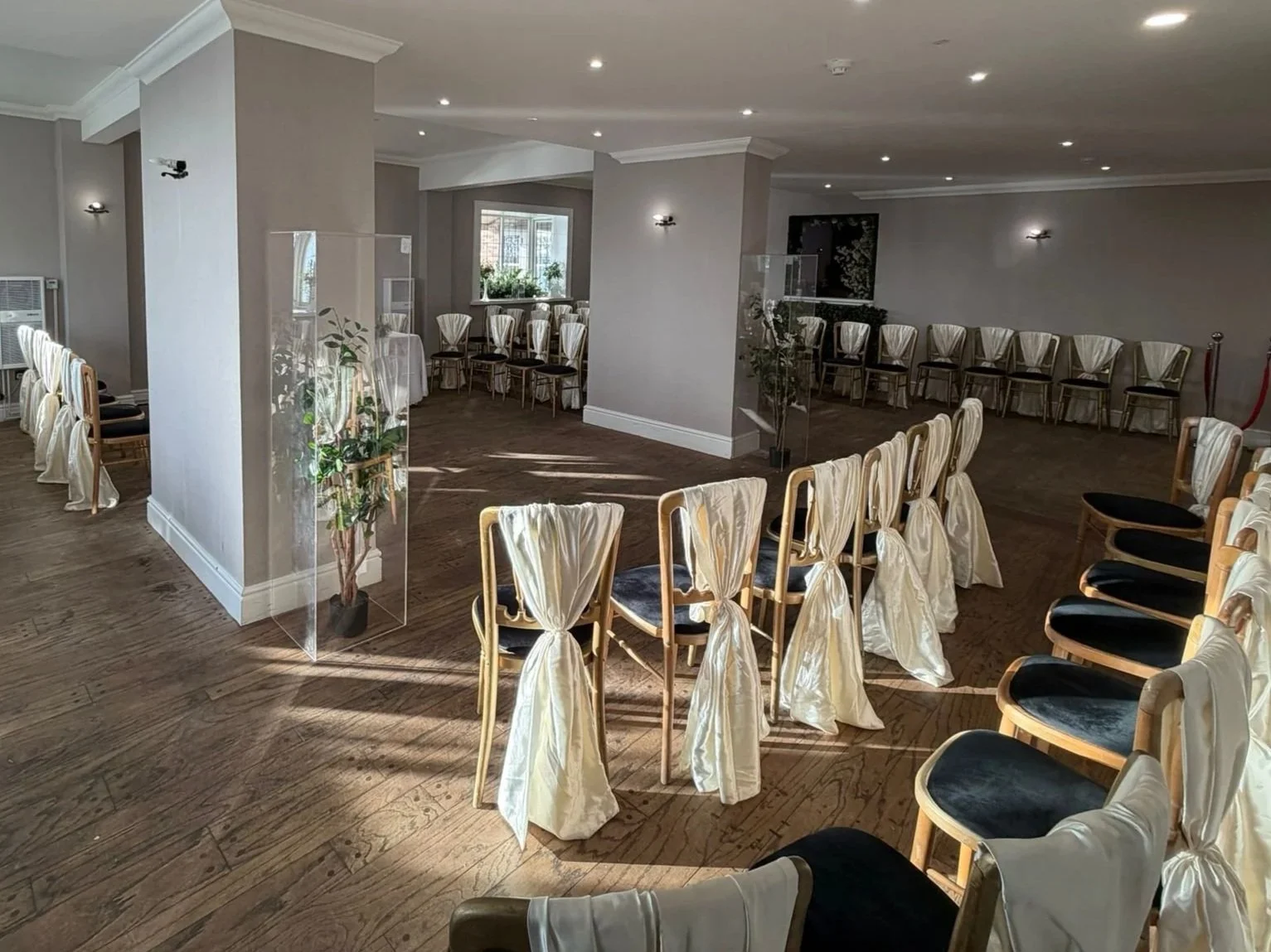 An empty event room with chairs decorated with white sashes, arranged in a semi-circle and rows, with plexiglass dividers and potted plants, ready for a ceremony.