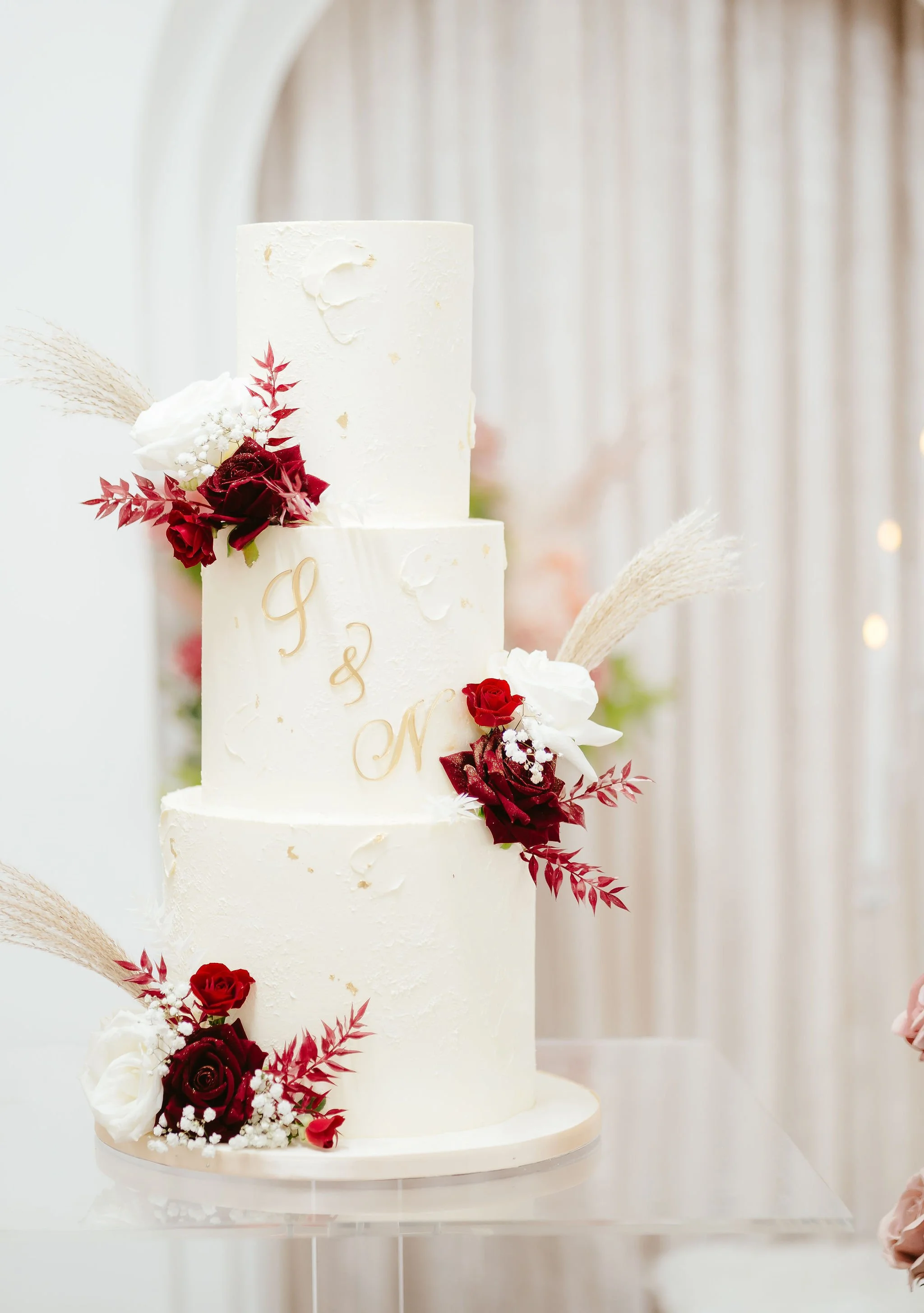 Elegant white tiered wedding cake decorated with red and white roses, baby's breath, and dried pampas grass, with gold lettering on the middle tier.