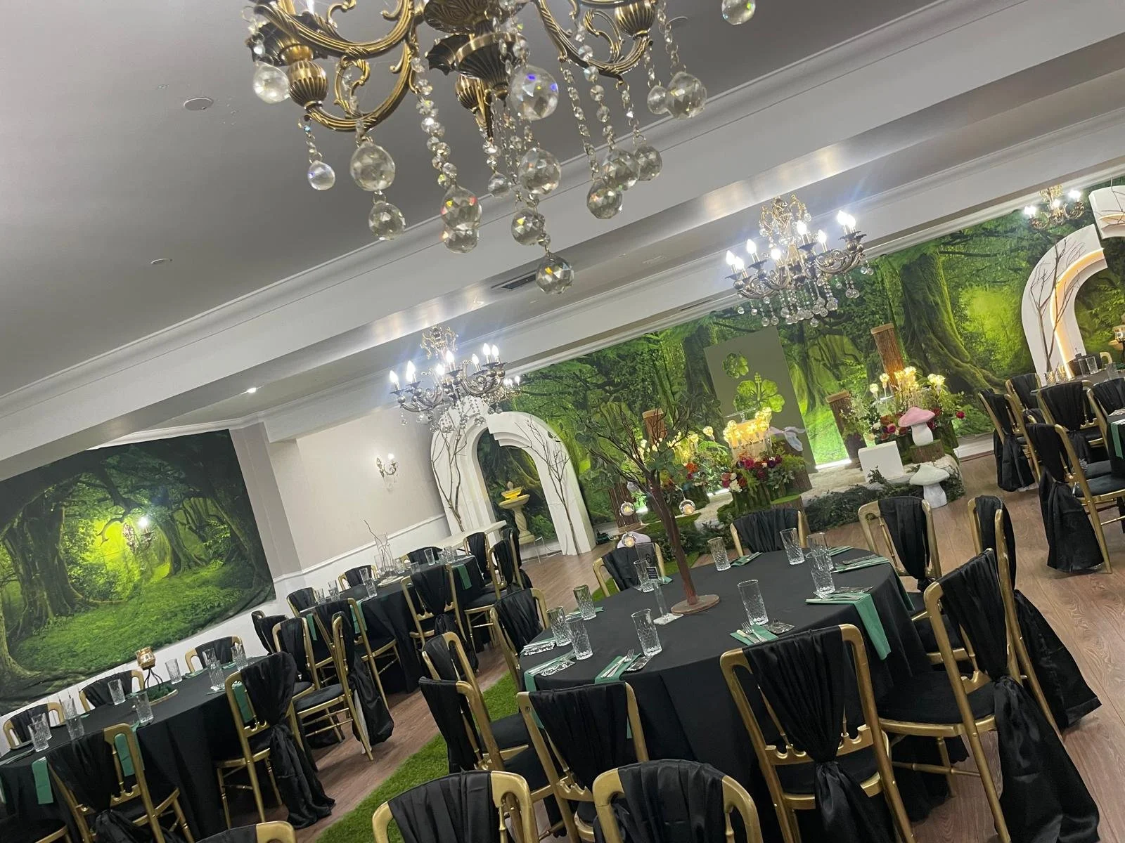 Decorated banquet hall with round tables covered in black tablecloths, black and gold chairs, chandeliers hanging from the ceiling, and a green forest-themed mural on the wall with floral arrangements and decorative lighting.
