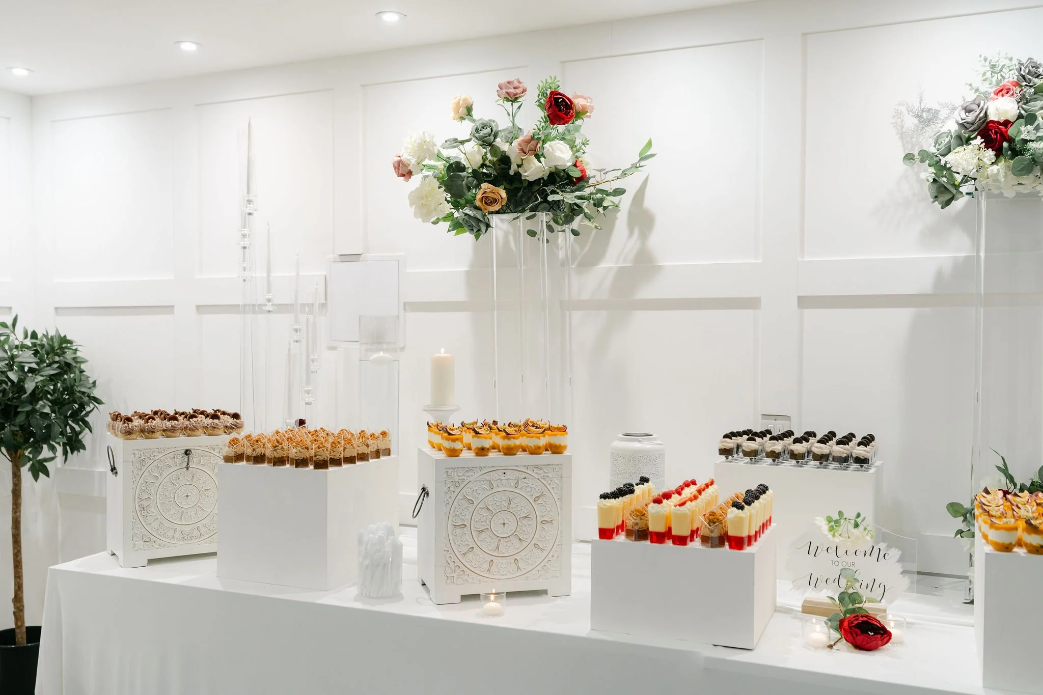 Dessert table with assorted mini desserts on white pedestals, decorated with large floral arrangements and a small sign that says 'Welcome to our wedding' in a white, modern setting.
