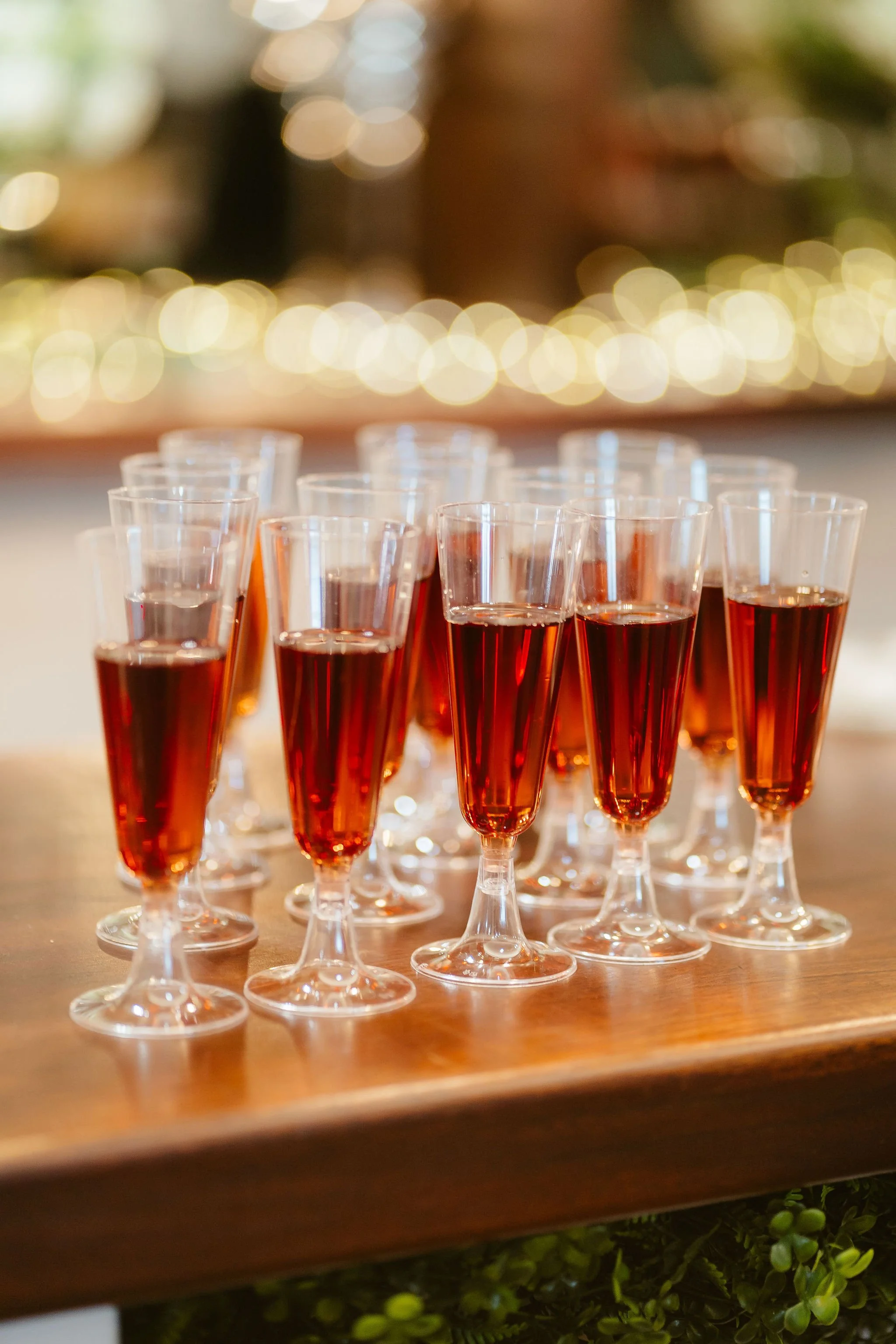 Multiple glasses of red or rose sparkling wine on a wooden table, with blurred fairy lights in the background.