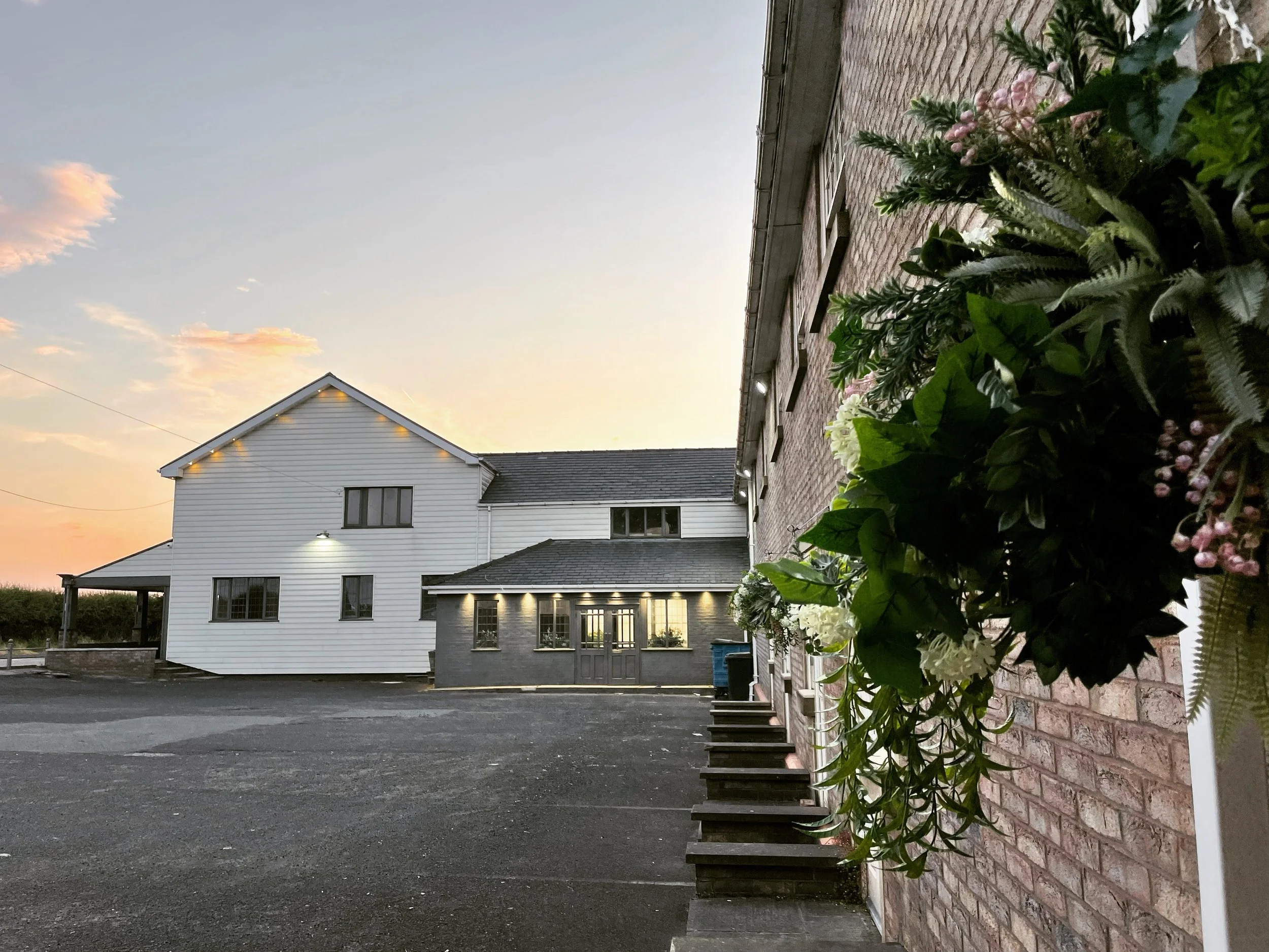 Exterior of a brick and white wooden building during sunset, with a sky showing orange and blue hues, a flower planter with green foliage and white and pink flowers on the right side.