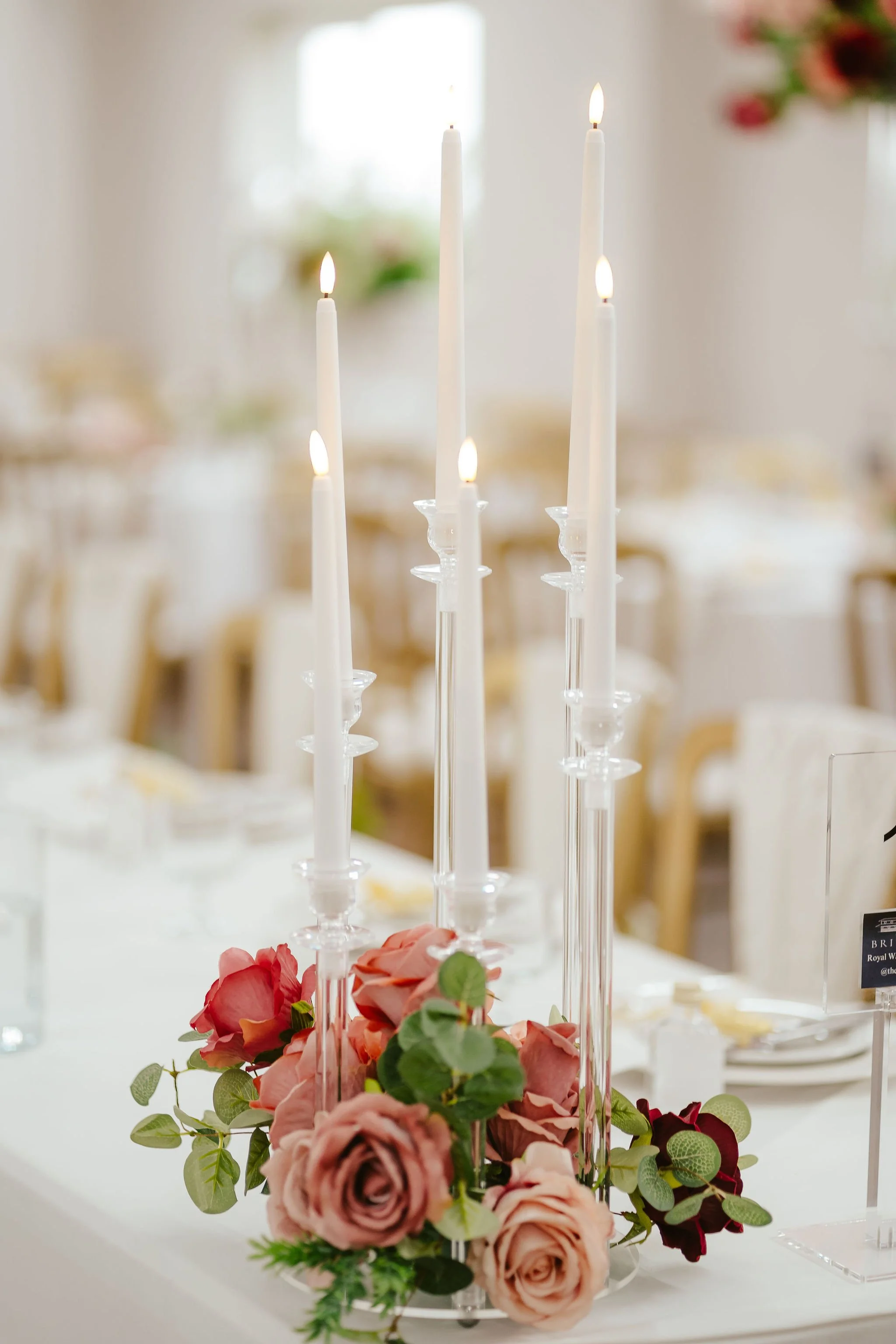 A table centerpiece featuring tall white candles in glass candle holders surrounded by a floral arrangement of pink and deep red roses with green leaves, set on a white tablecloth in an elegant venue.