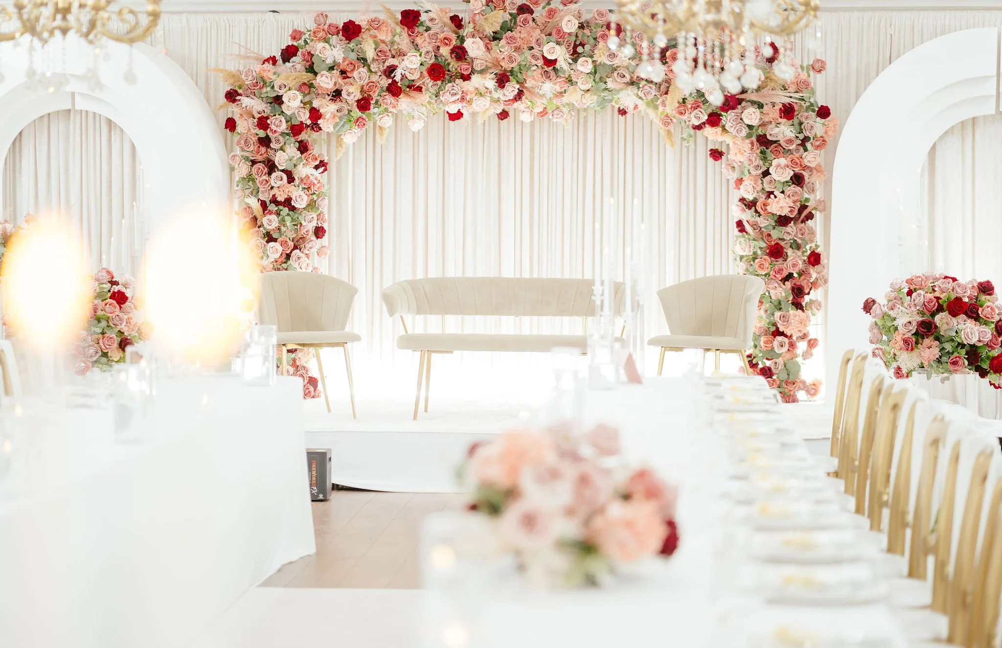 Elegant wedding reception decorated with pink and red roses on a floral arch, white drapes, and a stage with two white chairs and a sofa for the bride and groom, with a long table set for guests.