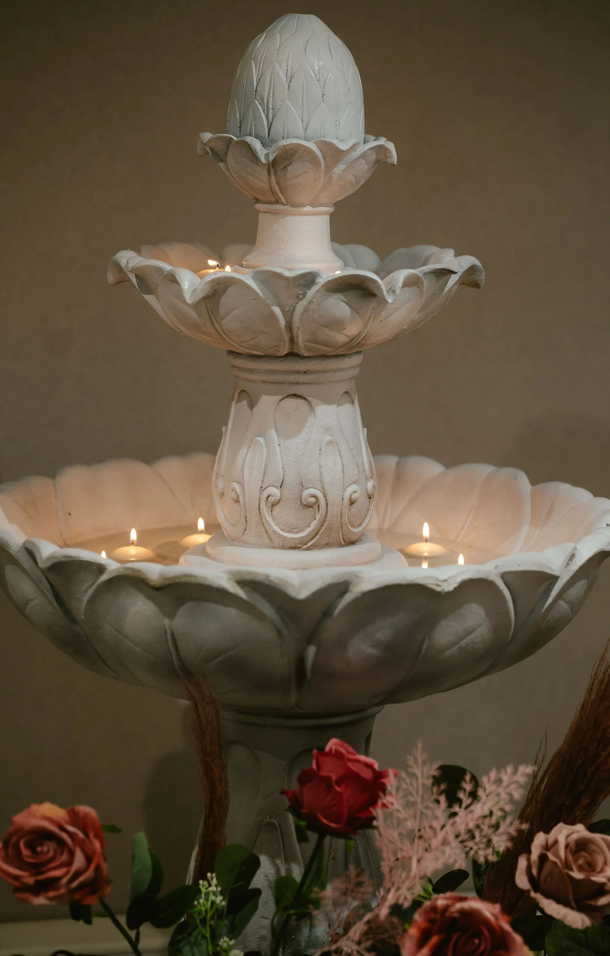 A decorative, multi-tiered white fountain with carved floral designs, surrounded by lit candles and flowers at the base.
