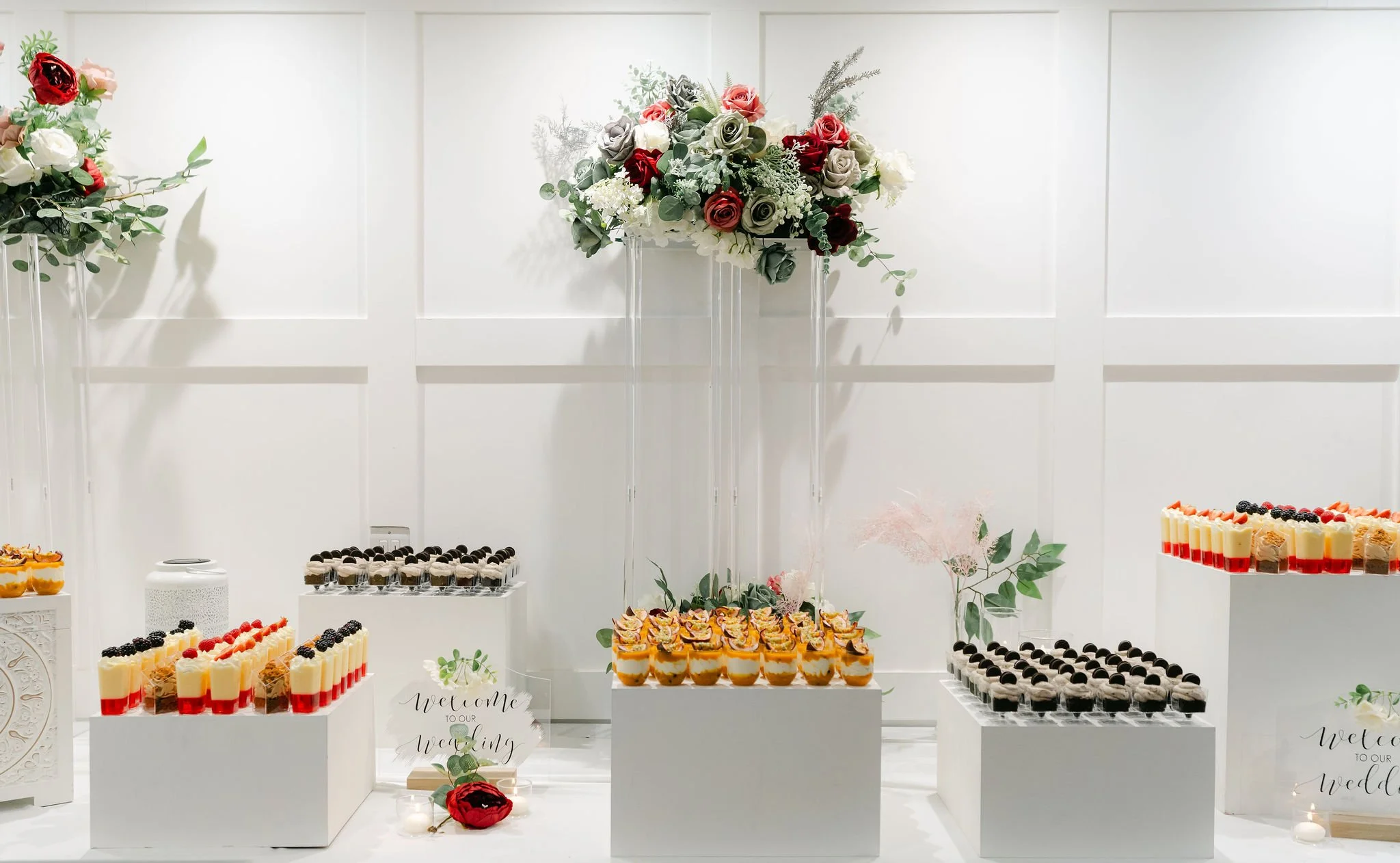 Elegant wedding dessert display with floral arrangements, assorted mini desserts, and welcome signs on white pedestals.
