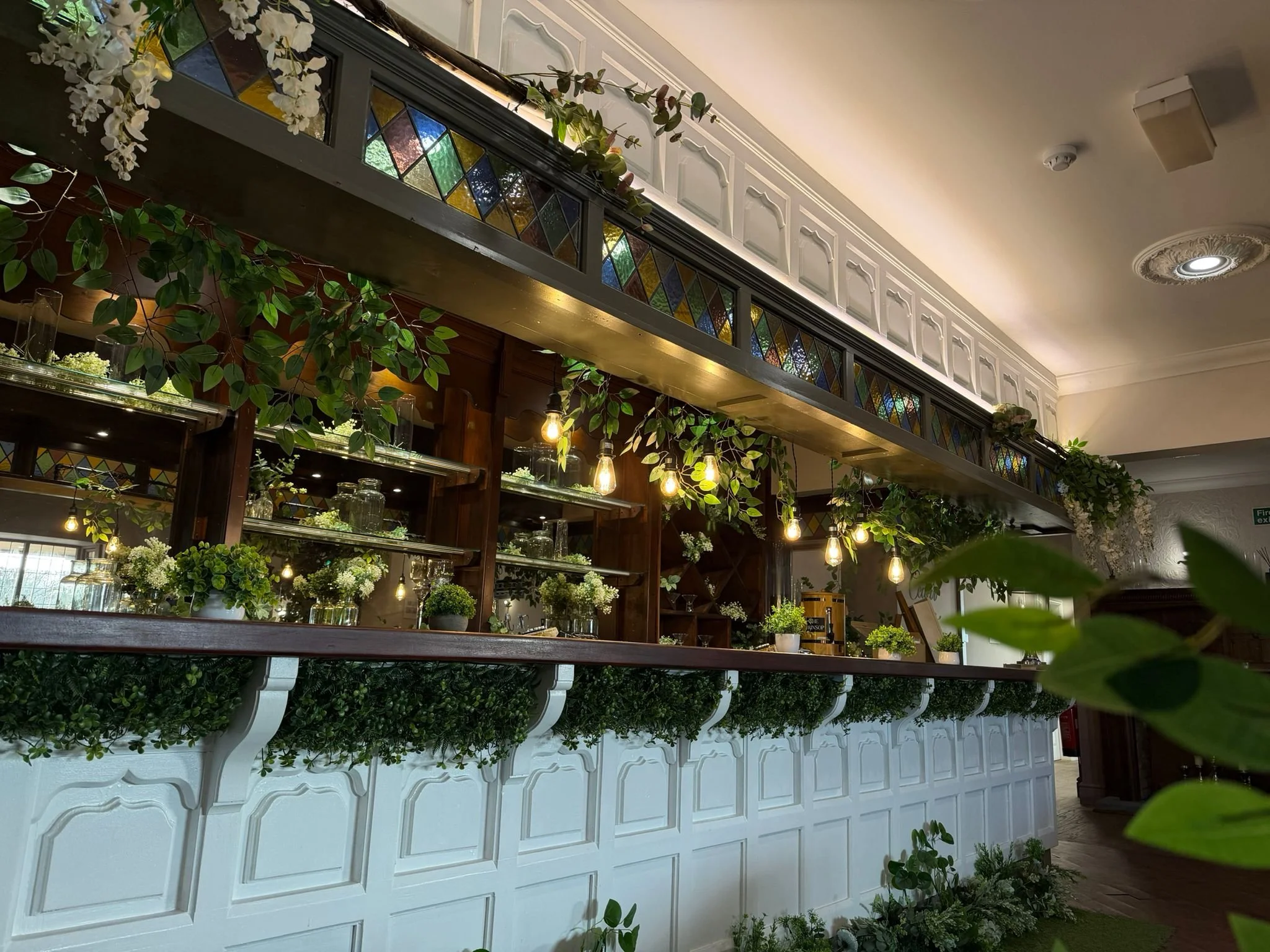 Interior of a decorated restaurant or cafe with a decorated bar, hanging Edison-style light bulbs, plants, and stained glass window panels.
