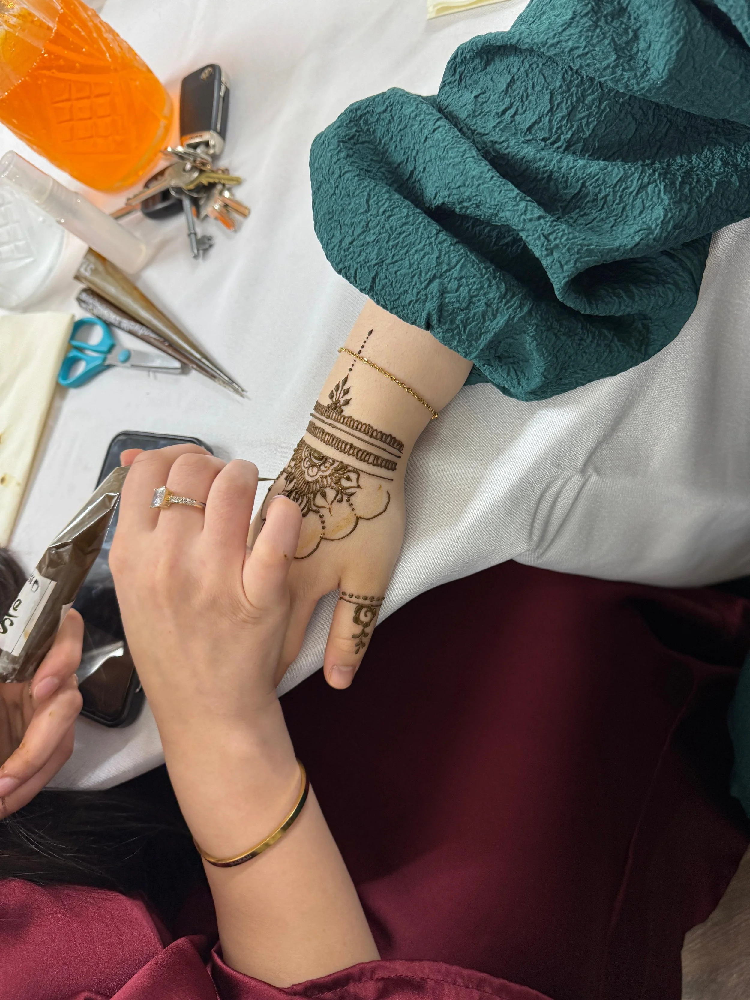 A person's hand with henna designs on the wrist and fingers, wearing a gold bracelet and ring, resting on a table. The table has items such as scissors, a phone, keys, a bottle, and other supplies. The person is wearing a teal sleeve.