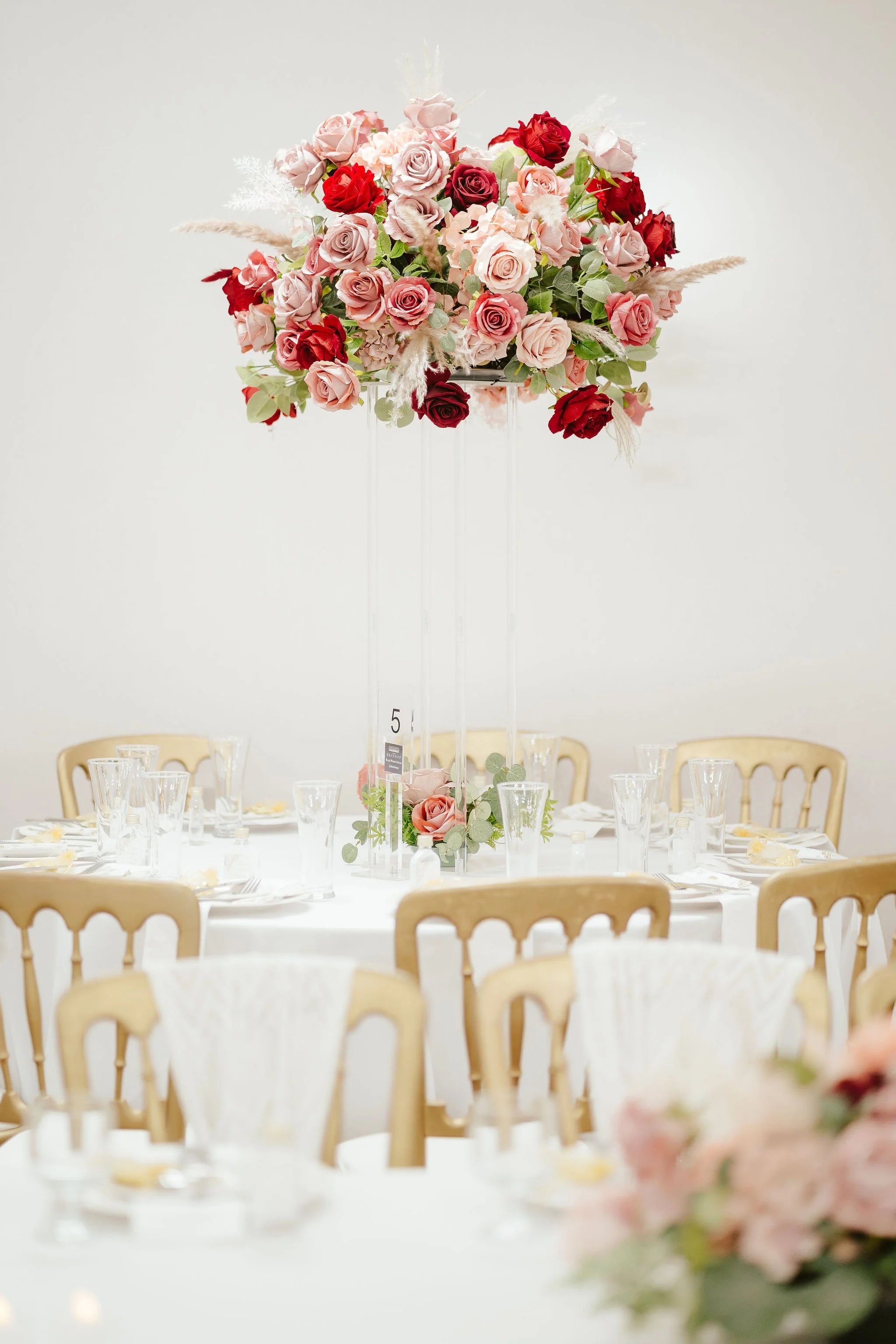 Elegant table setting with a large floral centerpiece of pink and red roses on a tall clear stand, surrounded by chairs and glassware for a formal event.