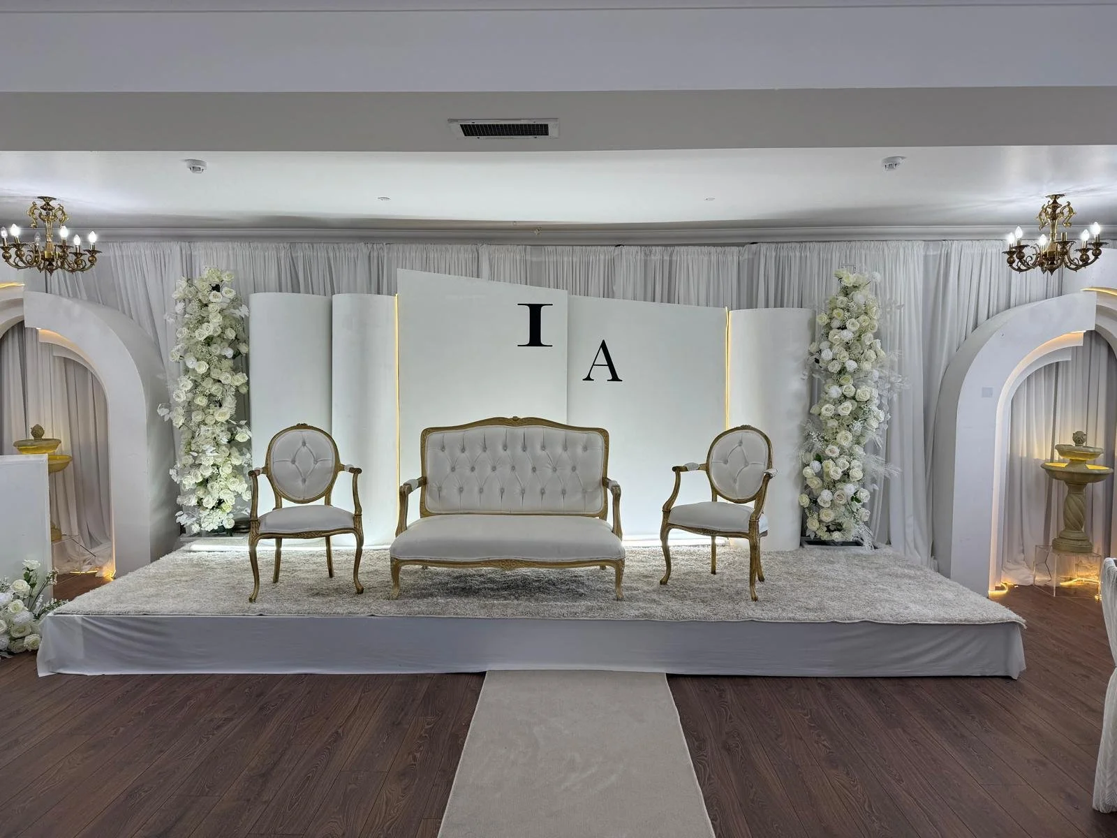 Elegant wedding stage decorated with white flowers, featuring a beige vintage-style sofa and two matching armchairs on a carpeted platform, with floral arrangements, golden accents, and white drapery in the background.